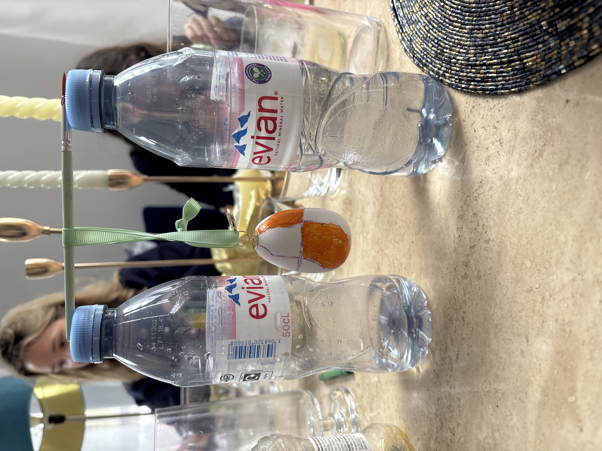 This indoor shot shows two Evian water bottles lying on a beige surface. One bottle is partially obscured by a painted egg with a green ribbon. A decorative candle holder with golden accents sits between the bottles. The image also captures parts of people in the background and other unidentified objects, including a straw hat. The location in London, United Kingdom, is not evident from the image.