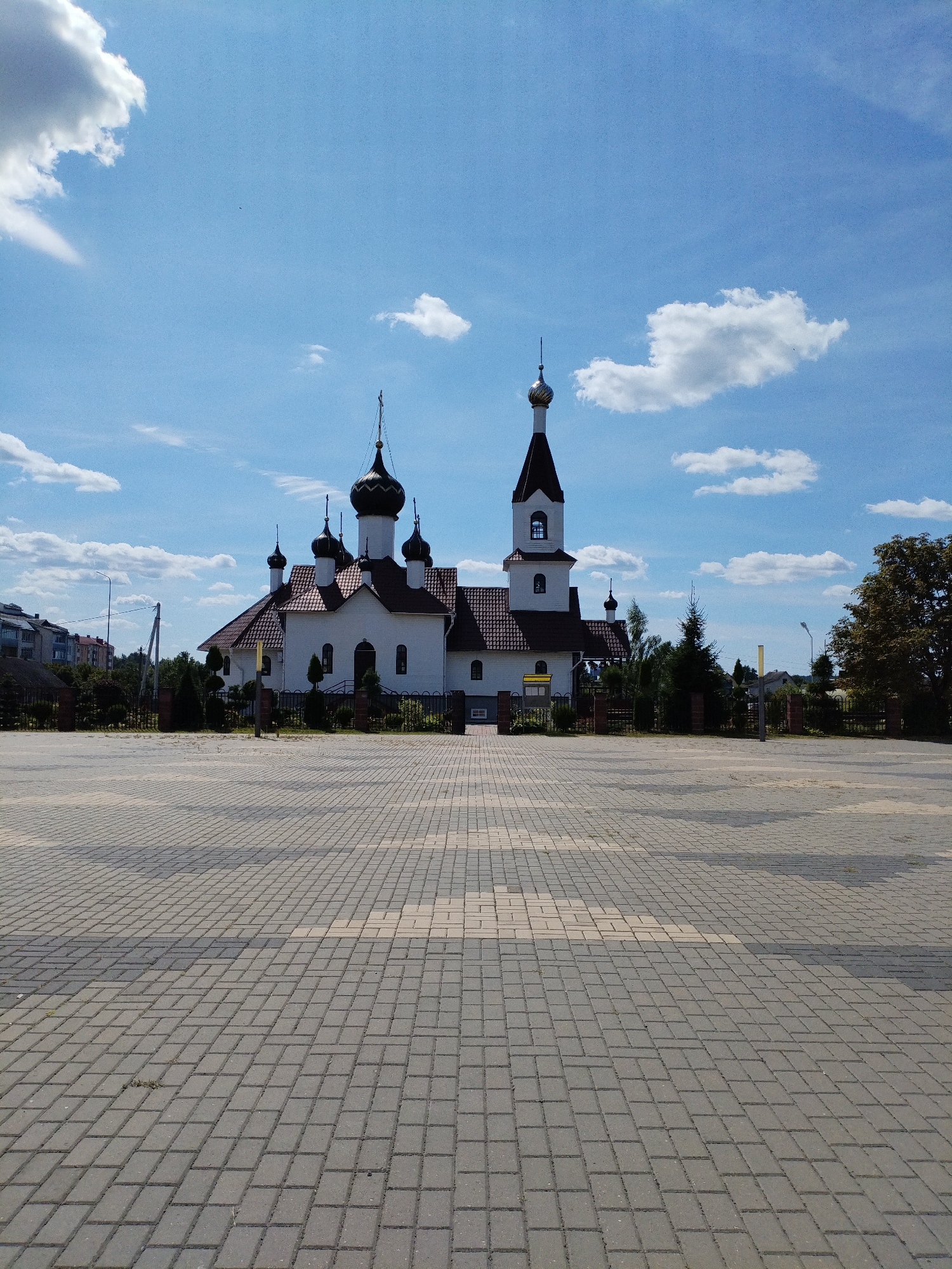 A low-angle, full outdoor shot captures a white church with a tall, dark-domed bell tower and several smaller dark domes, all topped with golden crosses, under a bright blue sky with scattered white clouds. The church features a red-tiled roof and dark arched windows. In front of the church is a vast, empty paved courtyard made of grey and beige interlocking bricks, with some shadows cast across it. To the left of the church, apartment buildings are visible, and to the right, trees frame the scene. The overall atmosphere is clear and sunny, suggesting a daytime scene in a tranquil, possibly residential area. The location is identified as a church in the city of Bialynichy, Belarus.