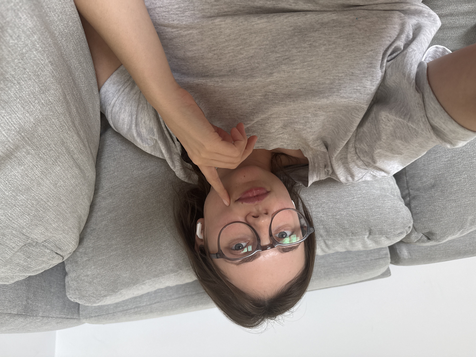 A young woman is lying on her side on a grey couch, with her head resting on a pillow. She is wearing a grey t-shirt and round, clear-rimmed glasses. She has on white wireless earbuds. Her right hand is raised, with her index finger touching her chin. Her eyes are open and looking directly at the camera. The lighting suggests it is daytime, and the setting appears to be indoors. The image is oriented with the woman's head at the bottom, suggesting it was taken as a selfie in a horizontal orientation.