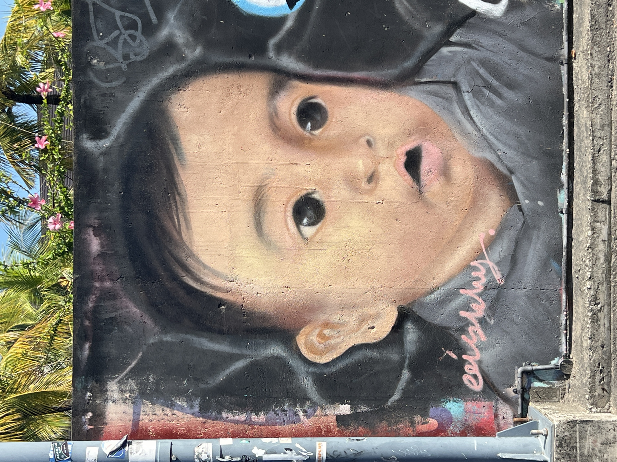 The image shows a close-up of a graffiti mural on a concrete wall, likely in the city of North Kuta, Indonesia. The mural depicts a lifelike portrait of a baby's face. The baby has large, dark eyes, a slightly open mouth, and light brown skin. The artwork is rendered with soft shading, giving the baby's skin a warm and luminous quality. The background of the mural is dark, with some abstract white graffiti lines. The baby is wearing a gray garment, possibly a shirt or onesie. Pink graffiti lettering is visible on the lower portion of the mural, beneath the baby's chin. Above the mural, the tops of palm trees and pink flowers are visible, suggesting an outdoor setting in a tropical or subtropical climate. A light gray metal pole, likely a street lamp or utility pole, is present on the left side of the image, with several stickers adhered to it. The bottom of the image shows a concrete ledge and steps, indicating the mural is at street level.