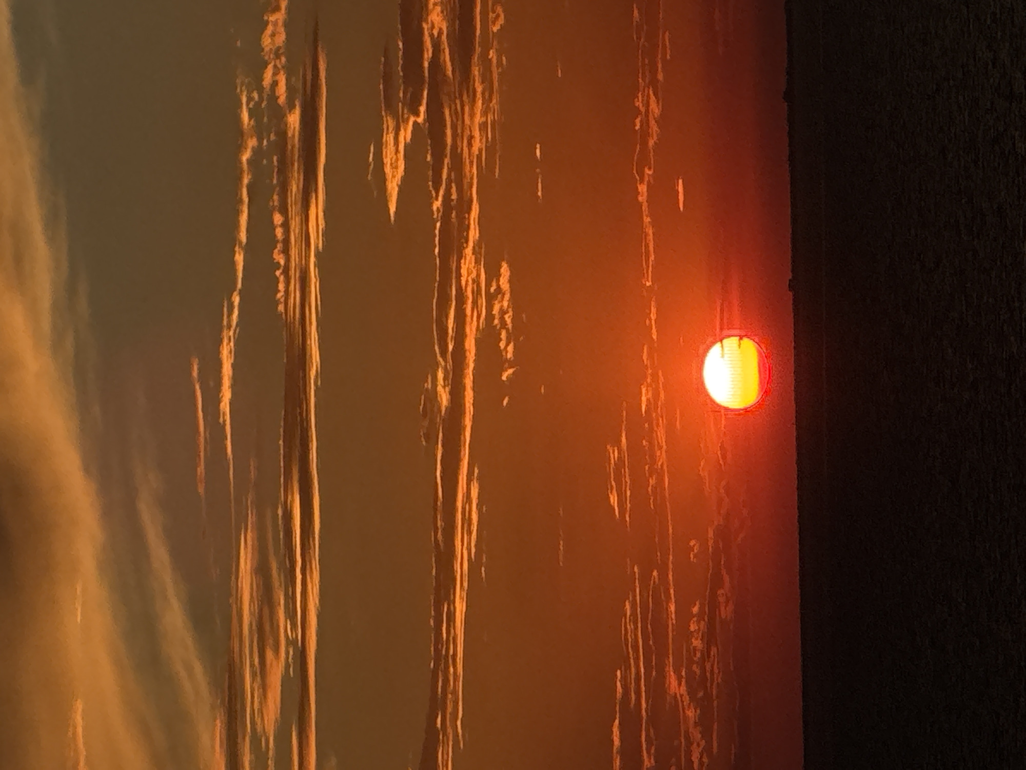 A sunset over a body of water creates a dramatic scene with vibrant orange and yellow hues. The sun, a bright orb with a yellow core, is partially obscured by clouds as it descends towards the horizon. Wispy clouds streak across the sky, catching the last rays of sunlight and intensifying the fiery colors. The lower portion of the image is dominated by a dark, silhouetted band, representing the water or a distant shoreline, meeting the luminous sky. The scene is devoid of any human presence or identifiable landmarks, suggesting a remote or natural setting. The overall impression is one of natural beauty and the end of a day.