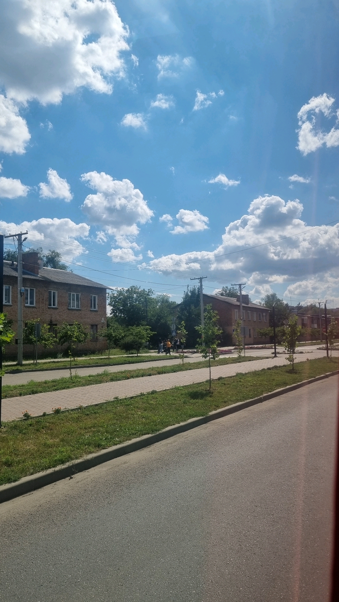 A view of a residential street in Smila, Ukraine, on a sunny day. The street is paved with asphalt and bordered by a sidewalk made of light-colored rectangular pavers. A strip of green grass with scattered weeds and a few small flowers separates the sidewalk from the street. On the left side of the street, there is a two-story brick building with a gray roof, multiple windows with white frames, and a visible chimney. Several young trees with green foliage are planted along the sidewalk in front of the building. Further down the street, a similar building is visible, also with a gray roof and red brick exterior. A group of people, appearing to be walking, are present on the sidewalk in the middle distance, near a crosswalk marked by traffic signs. Trees and utility poles line the street. The sky is a vibrant blue with numerous fluffy white clouds.