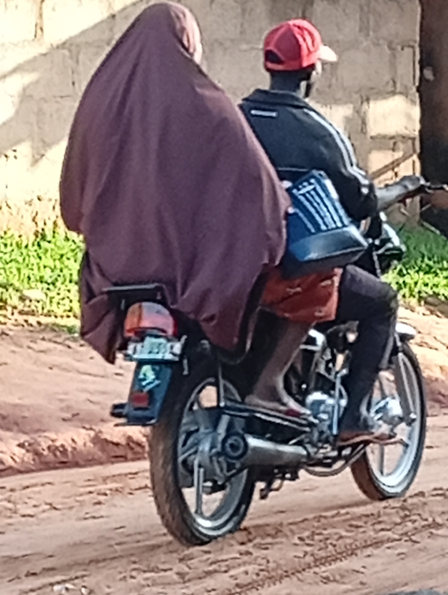 Two people, a driver wearing a red cap and a passenger covered in a dark purple cloth, are riding on a motorcycle on a dirt road in Tumfure, Nigeria. The driver is wearing a black jacket with white stripes on the sleeves and black pants. The passenger is seated behind the driver, with their body largely concealed by the large purple cloth. A blue bag with white vertical stripes is positioned between the driver and passenger, resting on the motorcycle. The motorcycle has a license plate with illegible characters. The background consists of a light-colored brick wall and green vegetation. The road surface is composed of sand and loose dirt.