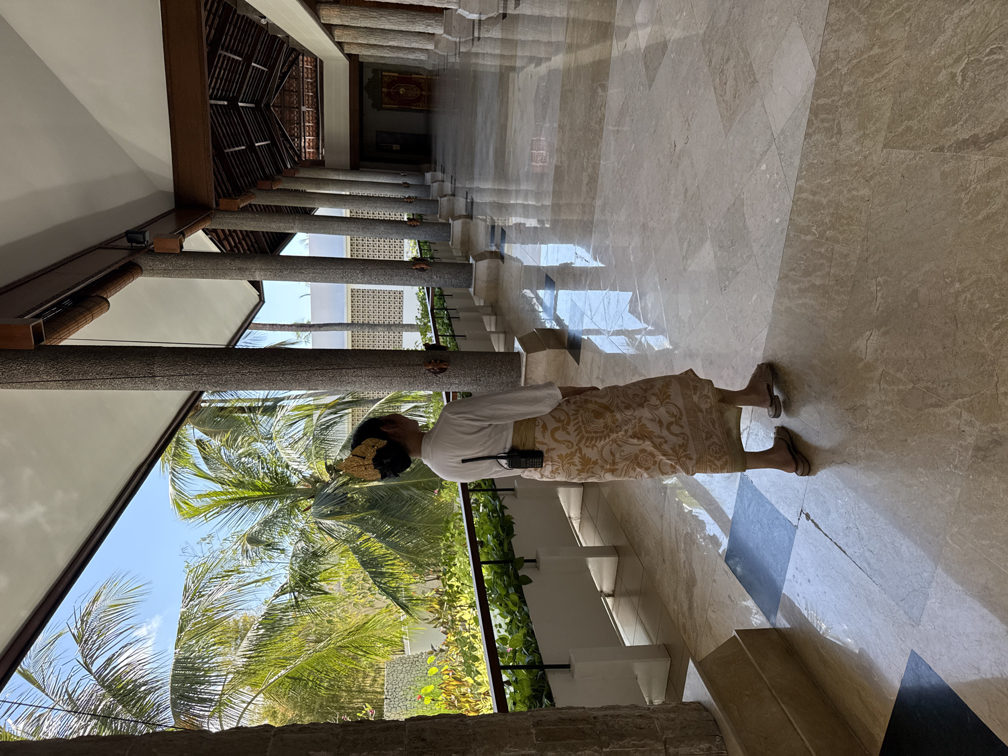 The image depicts a person standing in a spacious, open-air hallway or corridor, characteristic of traditional Balinese architecture, located in Kuta Selatan, Indonesia. The individual, viewed from behind, wears traditional attire consisting of a white long-sleeved top and a gold and beige patterned sarong. A gold ornamental piece adorns their dark hair, and a black walkie-talkie is clipped to their waist.
The hallway is lined with numerous grey, textured pillars supporting a dark wooden ceiling structure. The floor is made of highly polished, light-colored marble, which reflects the bright daylight, casting sharp, elongated shadows and mirroring the columns and ceiling. In the distant interior of the building, a red and gold tapestry or artwork is visible on a wall.
To the left of the columns, the scene opens to a vibrant tropical outdoors under a clear blue sky with scattered clouds. Prominent palm trees with their green fronds fill the foreground, alongside dense green foliage and bushes. A white, patterned wall or building exterior can be seen further back. A dark railing runs along the edge of the hallway overlooking this garden area.
The strong, defined shadows and reflections suggest bright sunshine, indicating that the time of day is likely midday or early afternoon. The setting evokes a sense of calm and tropical grandeur. There is no visible text in the image.