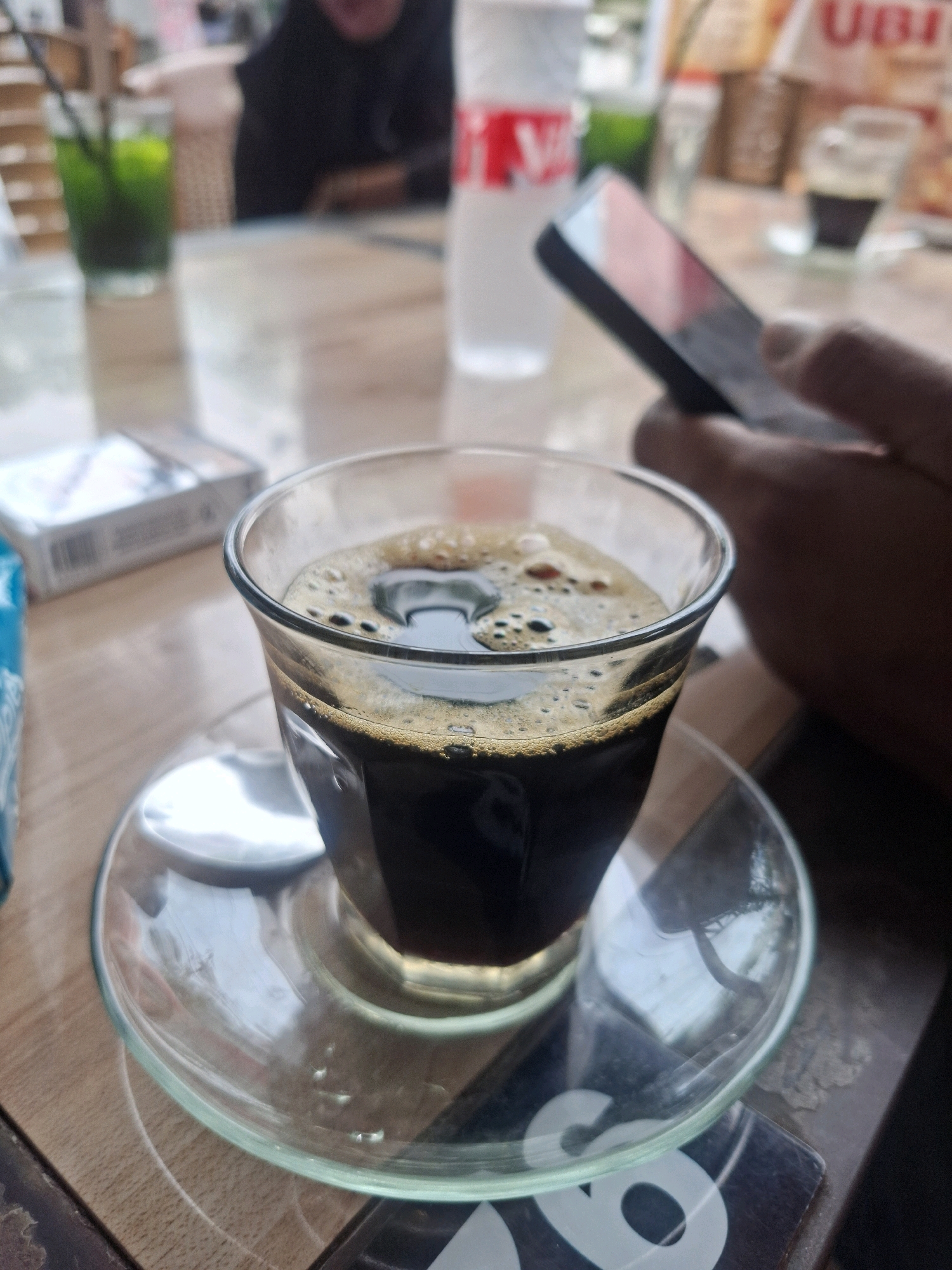 A close-up shot focuses on a clear glass filled with dark coffee, topped with a frothy crema, resting on a clear glass saucer. The saucer is on a light-colored wooden table. In the background, out of focus, a hand is holding a smartphone. To the left of the coffee cup, a pack of cigarettes is visible. Further back, a glass with a green beverage and a straw is on the table. The scene appears to be set in an outdoor cafe or restaurant. The context of "Banda Aceh, Indonesia" is not visually discernible from the image itself.