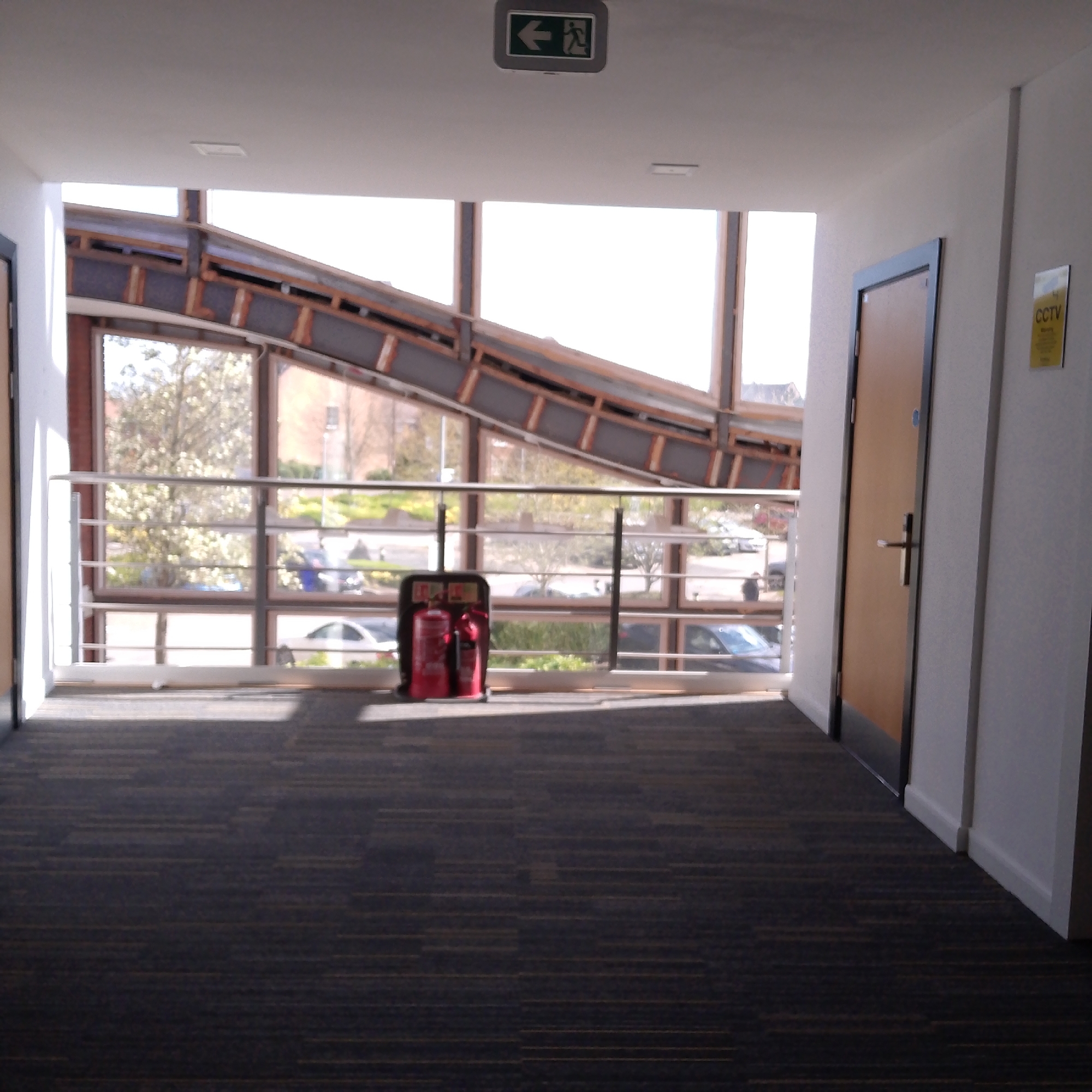 Here's a detailed description of the image:
**Scene:** An interior space, likely a corridor or walkway in a building. The space is illuminated by natural light.
**Location:** While not directly visible, given the presence of British signage (CCTV and the green 'exit' sign), the scene is most likely somewhere in the United Kingdom. Without more information, the exact location in Coventry cannot be determined.
**Objects:**
* **Exit Sign:** A green and white illuminated exit sign is mounted on the ceiling.
* **Fire Extinguishers:** A pair of red fire extinguishers are visible, secured in a black protective case.
* **Door:** A wooden door with a metal handle is positioned on the right side of the frame.
* **Window/View:** A large window with a metal railing offers a view of an exterior scene, including a building and greenery. A section of a metal and wood bridge structure is visible through the top panes of the window. Vehicles can be seen outside.
* **CCTV Sign:** A CCTV sign is affixed to the wall.
* **Carpet:** The floor is covered with a dark patterned carpet.
**Environment:**
* The location appears to be an indoor hallway area or connection way between buildings.
* There is natural light flooding the area.
**People:** No people are visible.
**Actions/Interactions:** There are no active actions or interactions occurring in the scene. The scene appears to be a static snapshot of a hallway.