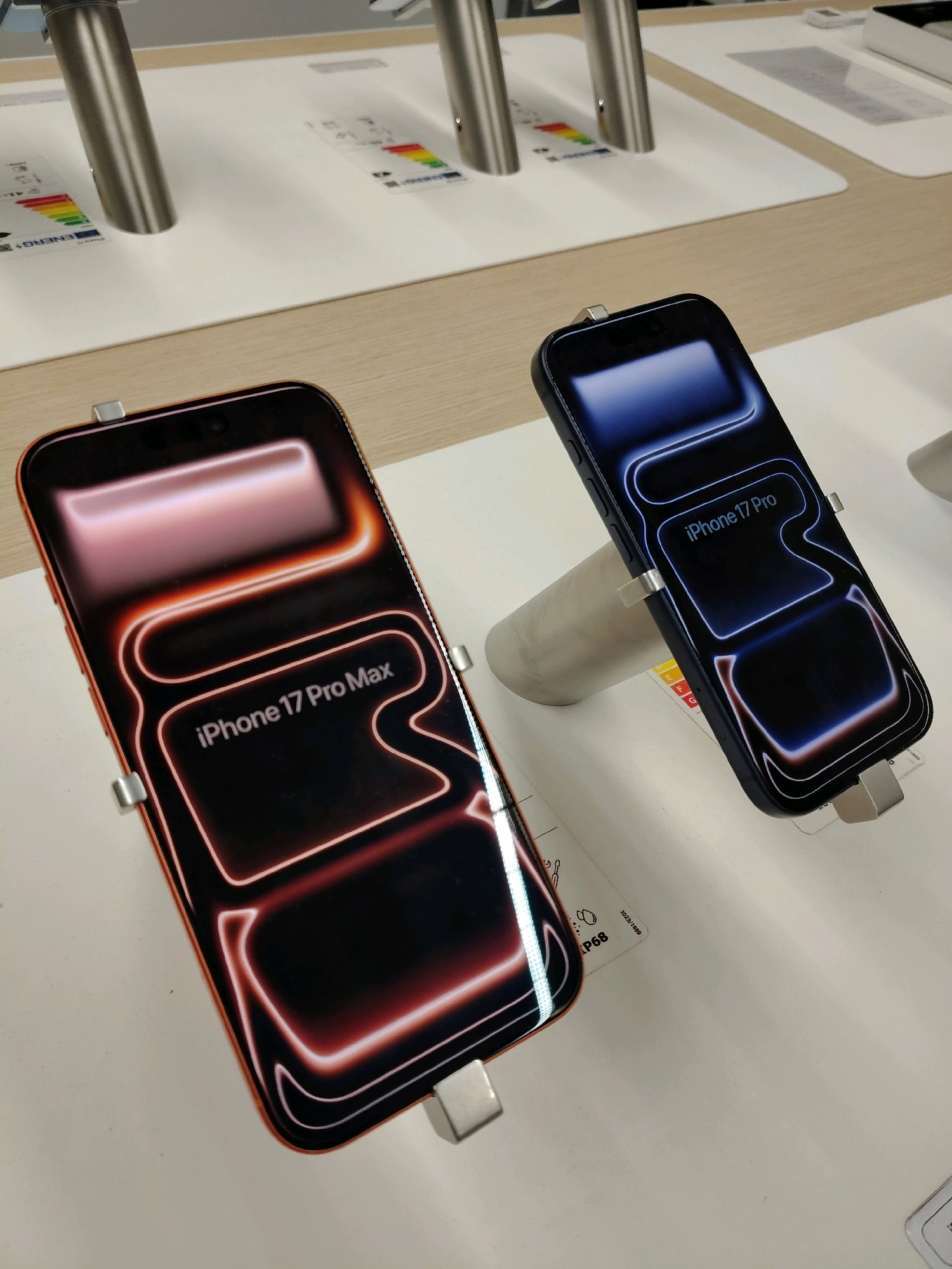 An eye-level, medium shot shows two iPhones displayed on a white counter. The iPhone on the left is orange and shows "iPhone 17 Pro Max" on its screen. The iPhone on the right is dark blue and displays "iPhone 17 Pro" on its screen. Both phones are propped up by silver stands. In the background, there are two metal poles and some energy efficiency labels on the white counter. The lighting is bright and even.