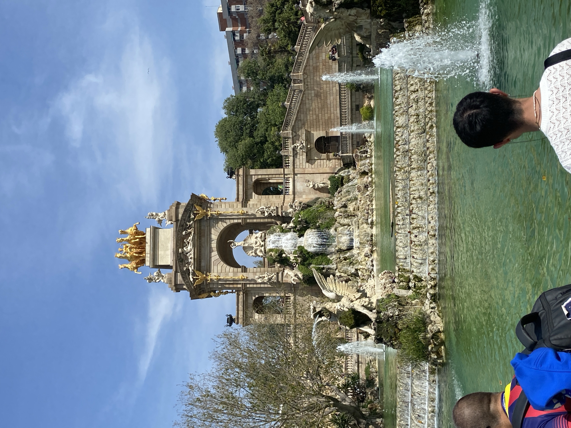 This image captures the majestic Cascada Monumental (Monumental Waterfall) in Parc de la Ciutadella, Barcelona, Spain, on a bright, sunny day. The grand fountain complex is the central subject, featuring multiple tiers, cascading waterfalls, and ornate architectural details.
At the very top of the fountain structure, a prominent golden quadriga (four-horse chariot) sculpture stands majestically, flanked by other golden figures. Below this, numerous white and stone statues of mythological figures, winged creatures, and other decorative elements adorn the various levels of the waterfall. Water dramatically spouts and cascades down several terraces, collecting in a large, greenish-blue pool at the base. Ornate arches and staircases are integrated into the fountain's design, with lush green foliage and trees providing natural accents around the stonework. In the upper left background, modern buildings are visible amidst more trees, indicating an urban park setting.
In the immediate foreground, to the right, two individuals are seen from behind, observing the fountain. One person wears a white, textured long-sleeved shirt and a beaded necklace. Another person, partially visible at the bottom right, wears a blue hat and a multi-colored shirt, with a black backpack resting beside them. Both appear to be tourists or visitors enjoying the impressive landmark.
The weather is clear and sunny, with a bright blue sky marked by a few wispy clouds. The strong sunlight casts clear shadows, suggesting it is midday or early afternoon. The scene conveys a relaxed and appreciative atmosphere as people take in the beauty and grandeur of the historic fountain. There is no visible text in the image.