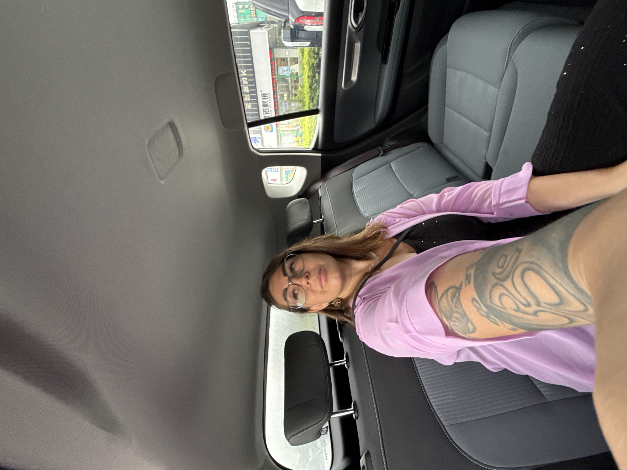 In Huizhou, China, a woman is seated in the back of a car, holding her phone to take a selfie. She is wearing glasses, a pink open-fronted jacket over a black top, and has a tattoo on her right arm. The car's interior features grey upholstery and a black headrest. Outside the window, businesses with Chinese signage and greenery are visible. The weather appears overcast.