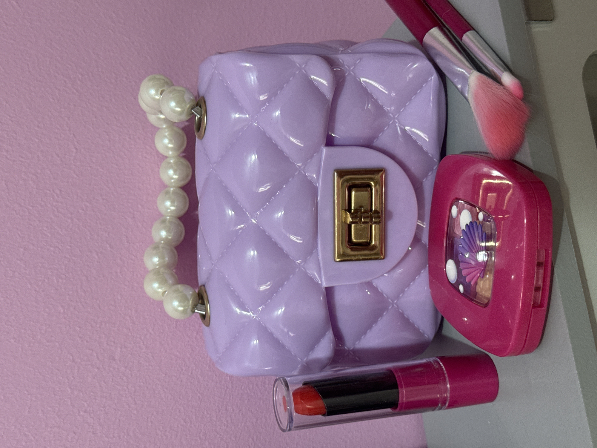 This media file is a close-up, overhead shot of a collection of children's play makeup and accessories arranged on a pink surface. The main subjects are a small, quilted, lavender-colored toy purse with a pearl handle, a bright pink compact with a seashell design, a tube of red lipstick, and two makeup brushes with pink bristles.
The purse is the central object, lying on its side. Its quilted texture is clearly visible, and it has a gold-colored clasp. Attached to the purse is a handle made of large, off-white faux pearls.
To the right of the purse is the pink compact. It's a rounded square shape with a clear window showcasing a colorful, glittery design that appears to be a seashell or fan. The compact is slightly open.
Below the purse and compact, a tube of red lipstick is positioned horizontally. The lipstick itself is a bright orange-red color, and it's partially extended from its clear plastic case.
Resting above the pink compact are two makeup brushes, likely for applying eyeshadow or blush. They have pink bristles that transition to a lighter shade at the tips, and silver ferrules connecting them to their dark handles.
The background is a soft pink, textured surface, suggesting a table or shelf. The lighting is bright and even, indicating an indoor setting, possibly a playroom or a dressing table. There are no people visible in the image. The overall impression is of a playful, curated arrangement of cosmetic toys. No text is visible in the image. The location context of Mikhaylovsk, Russia is noted but not visually evident in the image itself.