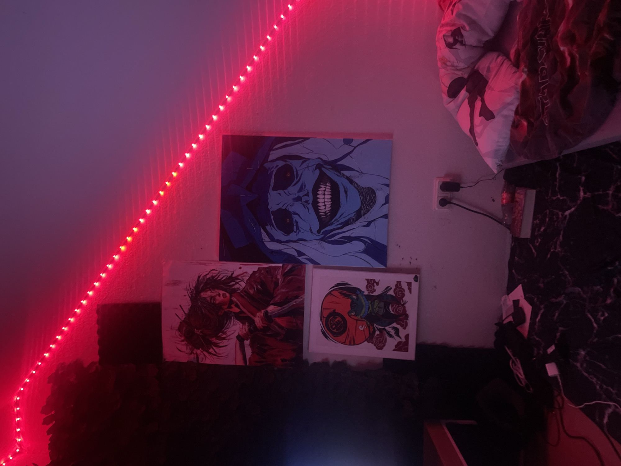 The image depicts a room interior. A wall is decorated with a string of red LED lights diagonally across the upper left portion. Three framed pictures are hung below the lights: one depicting a menacing blue figure with a wide grin, one showing a person with a sword, and one featuring a red and white design with a green character. Below the pictures is a dark, fuzzy material, possibly soundproofing foam. To the right of the pictures, a wall outlet with plugs is visible, along with a bed partially visible with anime-themed beddings. The environment appears to be lit with a mix of red and ambient lighting. The location cannot be determined from the image content.