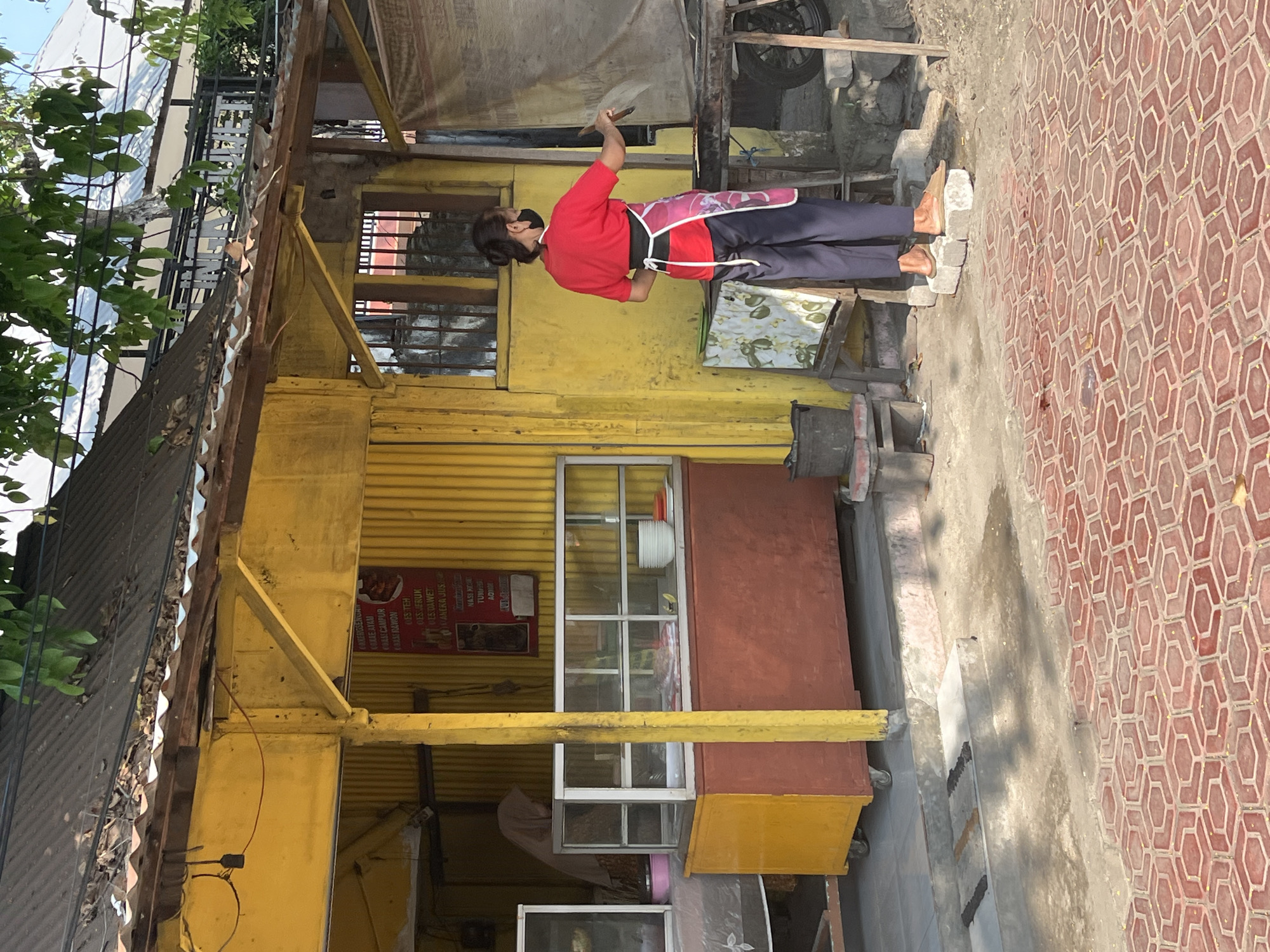 In Juwet Kenongo, Indonesia, a woman wearing a red t-shirt, a patterned apron, and dark pants is standing on two concrete blocks outside a yellow building. She is holding a cooking utensil and appears to be cooking or preparing food over a grill. Behind her, the building has a corrugated metal roof and a display case that holds bowls and other items. A sign with text is hanging on the wall. The ground in front of the building is paved with red hexagonal tiles. A person in a hijab is visible inside the building.