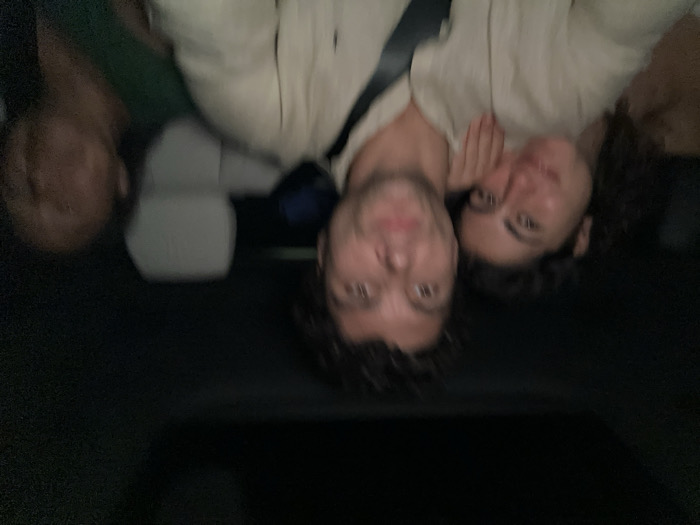 Three blurry people lie close in a dark interior. A man in a light button-down shirt has dark, curly hair and is partially obstructed by a black strap, possibly a seatbelt. To his right, a woman with curly hair smiles and touches her cheek. Another person is on the left, mostly out of focus and wearing a green shirt. They are likely in a car or similar vehicle.