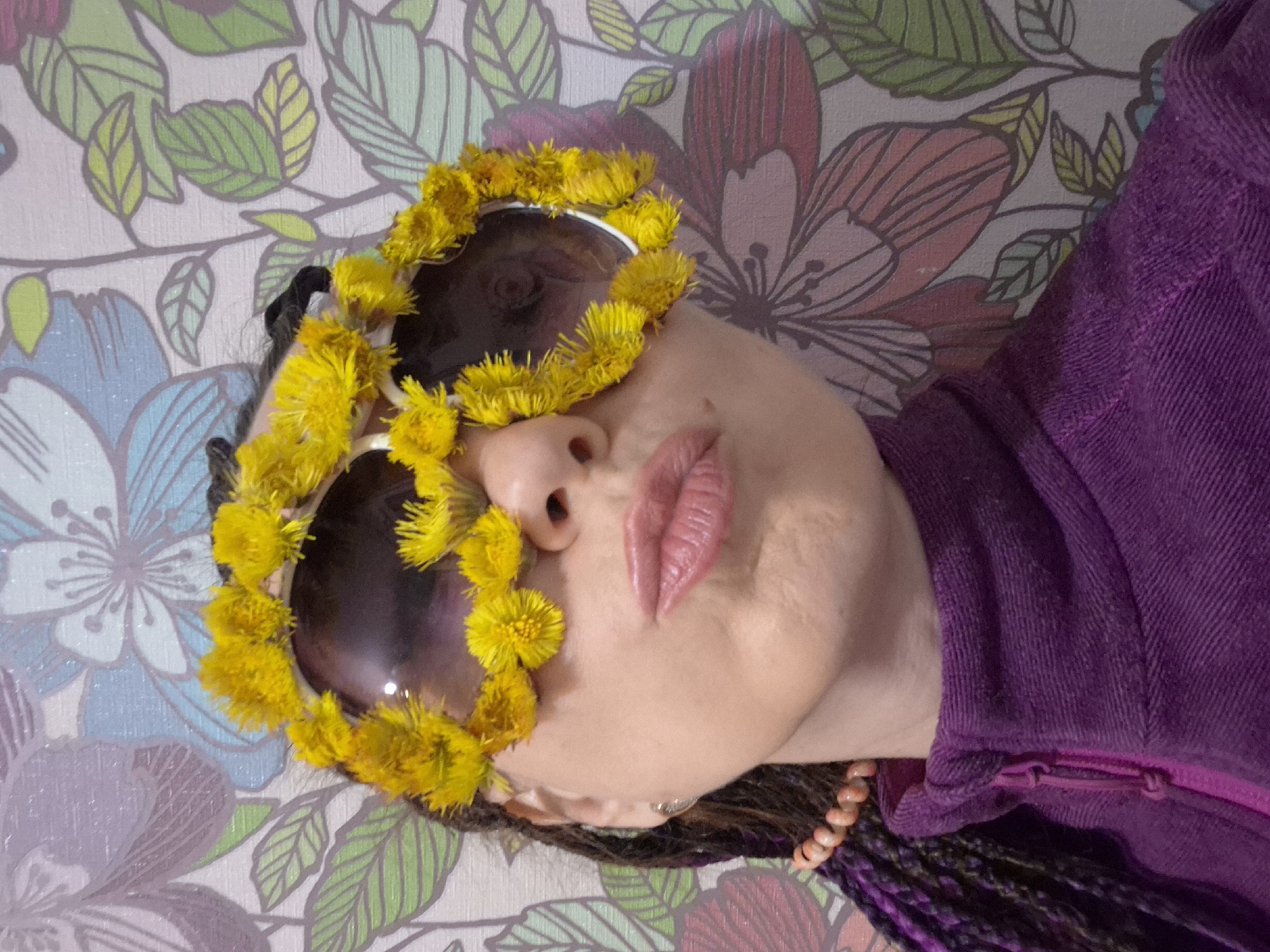 The image presents a close-up portrait of a person, with their head tilted to the right against a floral patterned background. The central focus is the person's face, partially obscured by large sunglasses that are elaborately decorated with numerous bright yellow flowers, possibly dandelions, around their frames. The person's lips are painted pink and are slightly parted, revealing a contemplative or posed expression.
They are wearing a purple garment, which appears to be a textured sweater or jacket with a visible zipper detail on the shoulder/collar area. Dark and purple braided hair is visible near their neck, along with a string of light-colored beads, suggesting a necklace or bracelet. The background is a wallpaper featuring a pattern of large, stylized flowers and leaves in shades of blue, green, pink, and white. The scene appears to be set indoors, under soft, diffused lighting. There is no specific activity taking place other than posing for the photograph, and no visible text.