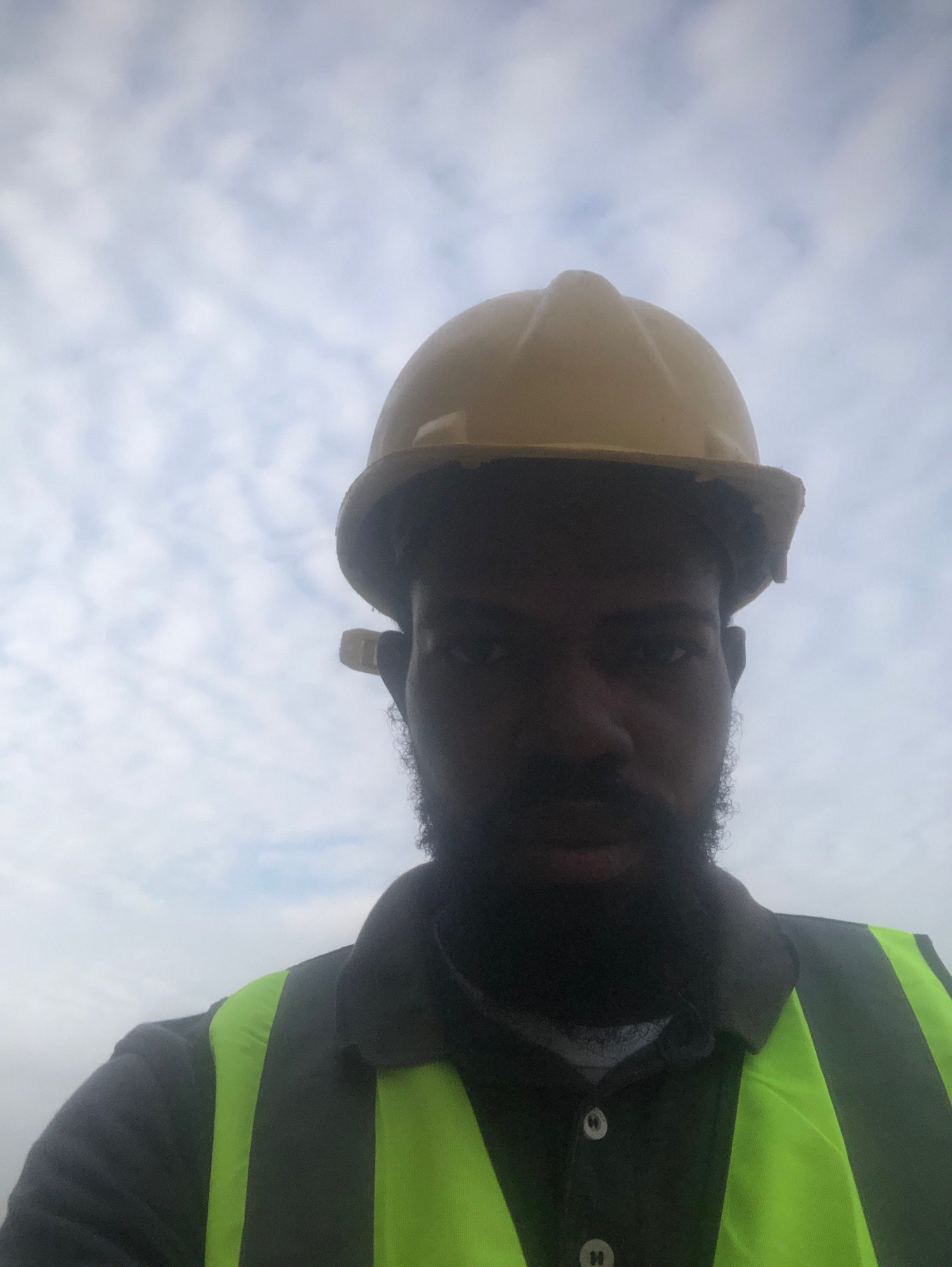 A medium shot selfie captures a Black man wearing a yellow hard hat and a high-visibility safety vest over a dark collared shirt. He has a full beard and is looking directly at the camera. The background is an overcast sky with scattered clouds. The image appears to have been taken outdoors, with no specific landmarks to indicate the location in Ikeja, Nigeria.
