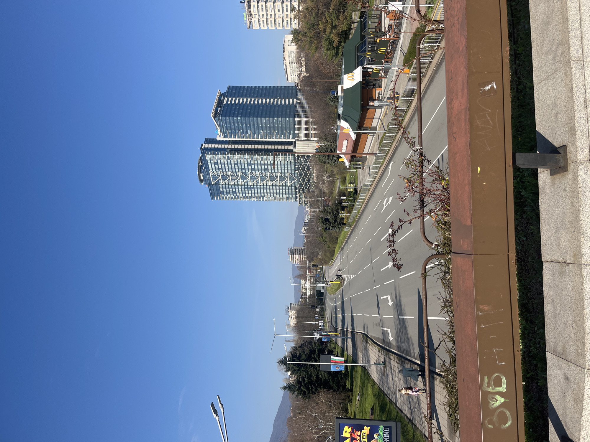 The image shows a view of a wide avenue in Sofia, Bulgaria, from an elevated position, likely a bridge or overpass. The avenue is divided into multiple lanes with white lane markings and directional arrows painted on the asphalt. Buildings of varying heights line the avenue, including modern high-rise buildings and older, shorter structures. A McDonald's restaurant is visible on one side of the street. Along the pedestrian walkway, there are flags and lamp posts. A brown metal railing with graffiti is in the foreground on the right, obscuring some of the view. The sky is clear and blue, suggesting a sunny day. In the distance, mountains can be seen. A few people are walking along the sidewalk.