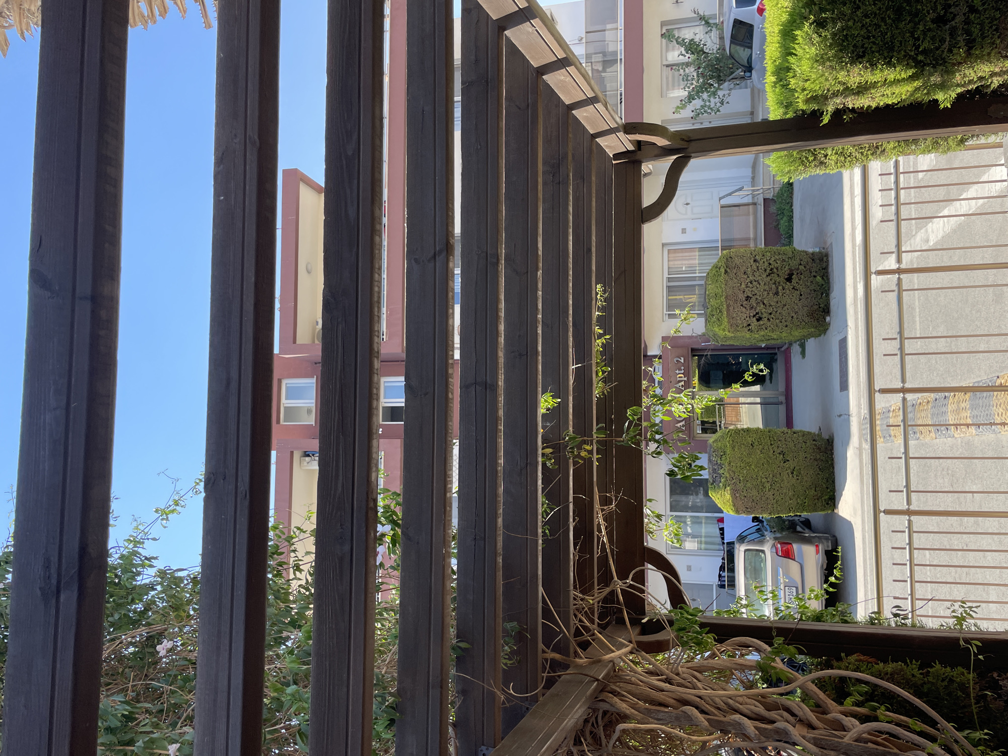 The image shows a view looking through a wooden pergola with horizontal beams. Vegetation, including vines and green leaves, is growing on and around the pergola. In the background, there is a multi-story building with a red upper section and a white lower section. The lower section of the building has multiple windows and doorways. Two rectangular, trimmed green bushes are situated in front of the building. A light-colored sedan is parked to the left, partially obscured by the bushes. A sign with "Apt. 2" is visible on the building. A metal fence with vertical bars and yellow and black striped speed bumps is in the foreground. The sky is clear and blue. The scene appears to be an outdoor area of an apartment complex or residential building, likely in a Mediterranean climate given the vegetation and architecture.