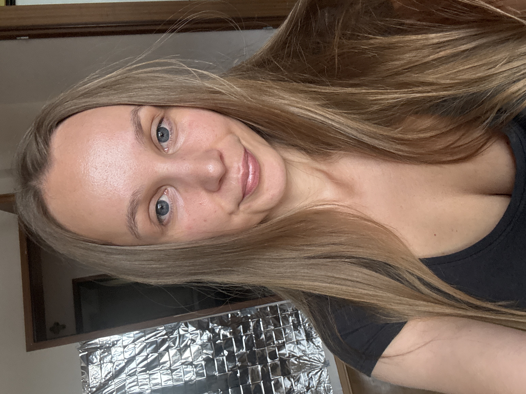 A young woman with long, straight, blonde hair is pictured in a close-up, head-and-shoulders shot. She is wearing a black t-shirt. Her face is turned slightly to the side, and she is looking at the camera with blue eyes. Her expression is a gentle smile. In the background, there is a door and a wall covered in what appears to be a metallic, reflective material. The lighting suggests an indoor setting, possibly a room in a residential property. There is no visual information in the image that can be used to determine the specific location in Überherrn, Germany.