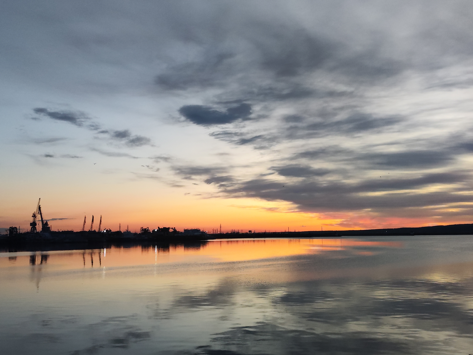 The image depicts a sunset over the Kama River in Chaikovsky, Russia. Silhouetted against the orange sky are industrial cranes and other equipment, likely related to shipbuilding or a port facility. The water surface reflects the sky and the silhouetted shoreline. The sky above the horizon shows a transition from orange hues near the horizon to gray and blue hues higher up, with scattered clouds. The water is relatively calm, producing a smooth reflection of the sky and shoreline.