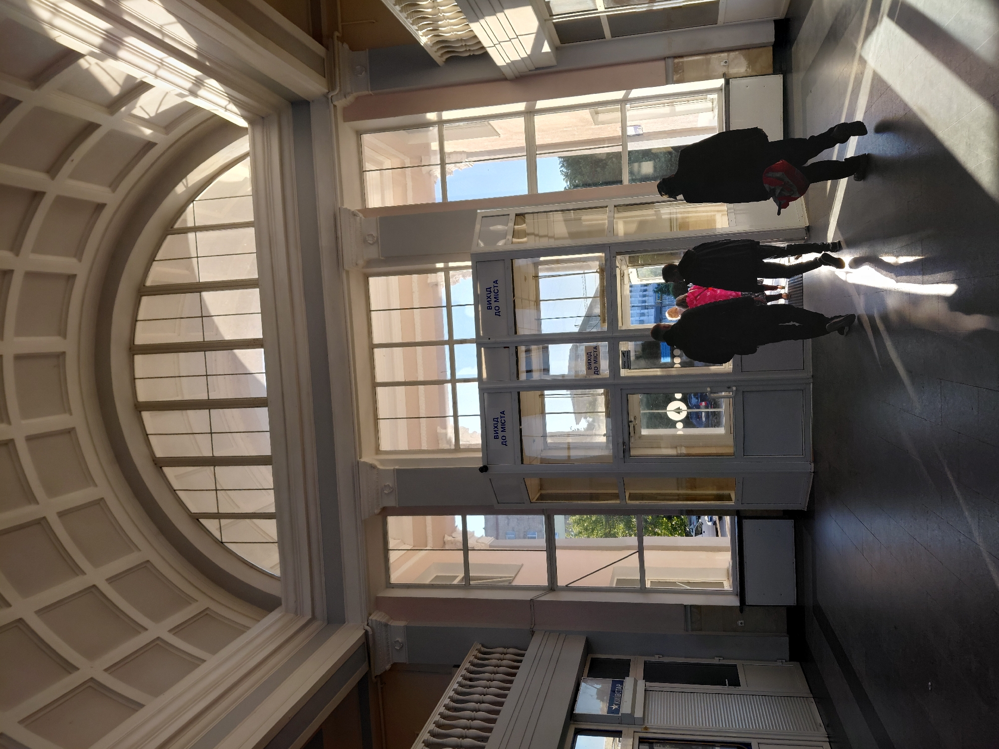 An indoor, well-lit scene shows a grand hall or passageway, possibly within a train station or public building, with several people walking towards an exit. The image is taken in bright daylight, casting long, distinct shadows across the floor from the people and architectural elements, suggesting either morning or late afternoon sun.
The most prominent feature is a large, arched window structure on the left, with numerous smaller rectangular panes and a decorative, coffered ceiling-like pattern above it. To the right, a wall of vertical rectangular windows offers a clear view to the outside, revealing glimpses of trees and other buildings under a clear sky. A classical-style balustrade is visible towards the bottom right, adding to the architectural grandeur.
Several figures, mostly seen as dark silhouettes against the bright light from the windows, are moving towards two glass doors that serve as an exit. One person in the foreground wears a red backpack. Signs affixed to the glass doors clearly display "ВИХІД ДО МІСТА" (Vykhid do mista), which translates from Ukrainian as "Exit to the City," indicating the purpose of this area. Further down on the right, another sign with the text "КИЇВСТАР" (Kyivstar) is visible, identifying a Ukrainian mobile communications company, suggesting the location is in Ukraine. The floor is dark and reflective, accentuating the strong light and shadows.