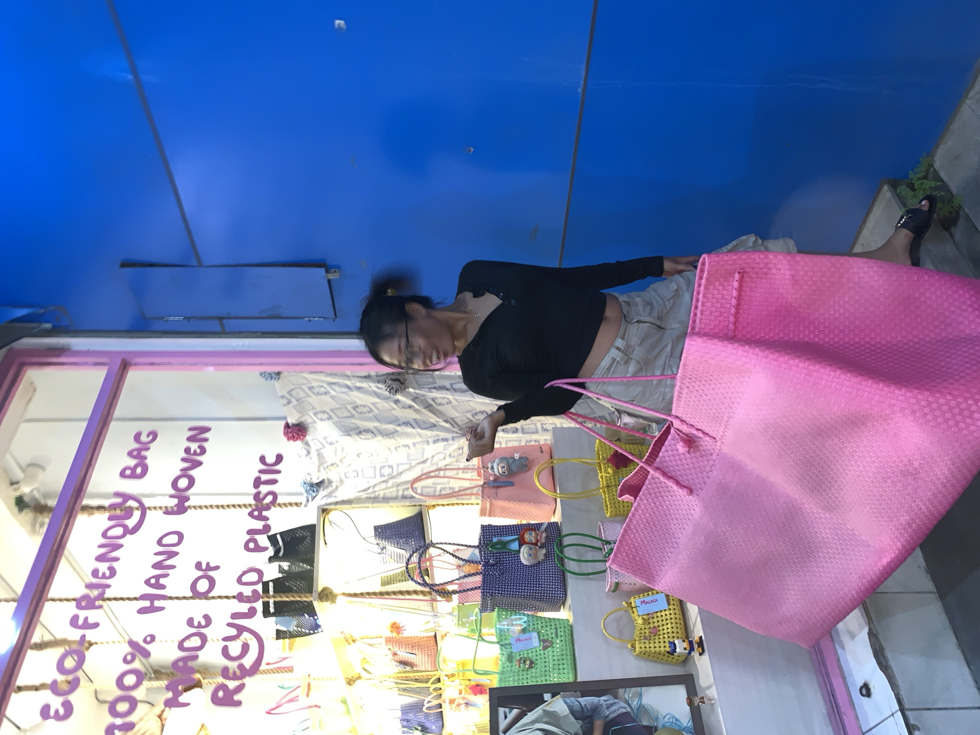 A medium, indoor shot captures a woman wearing a black, long-sleeved shirt and beige pants, examining a display of woven bags in what appears to be a store window. She is also carrying a large pink bag. The bags on display come in various colors, including green, yellow, blue, and pink, with small embellishments attached. A sign is visible on the window, reading "100% ECO-FRIENDLY BAG. HAND WOVEN MADE OF RECYCLED PLASTIC". The shot seems to be taken in Ubud District, Indonesia, based on the merchandise and the environment.