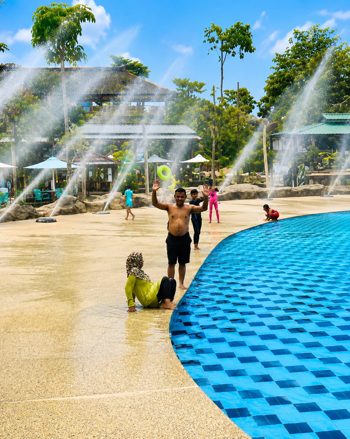 In the city of Eampat Kwalamencirim, Indonesia, a water park scene unfolds under a bright blue sky dotted with fluffy white clouds. Several fountains spray water into the air, creating a misty atmosphere. In the foreground, a woman wearing a lime green long-sleeved shirt and black pants, along with a patterned headscarf, sits on the edge of a bright blue tiled swimming pool, her legs submerged in the water. To her right, a man with bare chest and black shorts stands with his arms raised, appearing to wave. Behind him, two children, one in a black swimsuit and the other in a pink swimsuit, are also present. Further in the background, another child in a red swimsuit is visible near the pool. The ground surrounding the pool is a light beige, speckled surface, with scattered rocks and vegetation. In the distance, the water park features covered seating areas with thatched roofs and tall green trees.