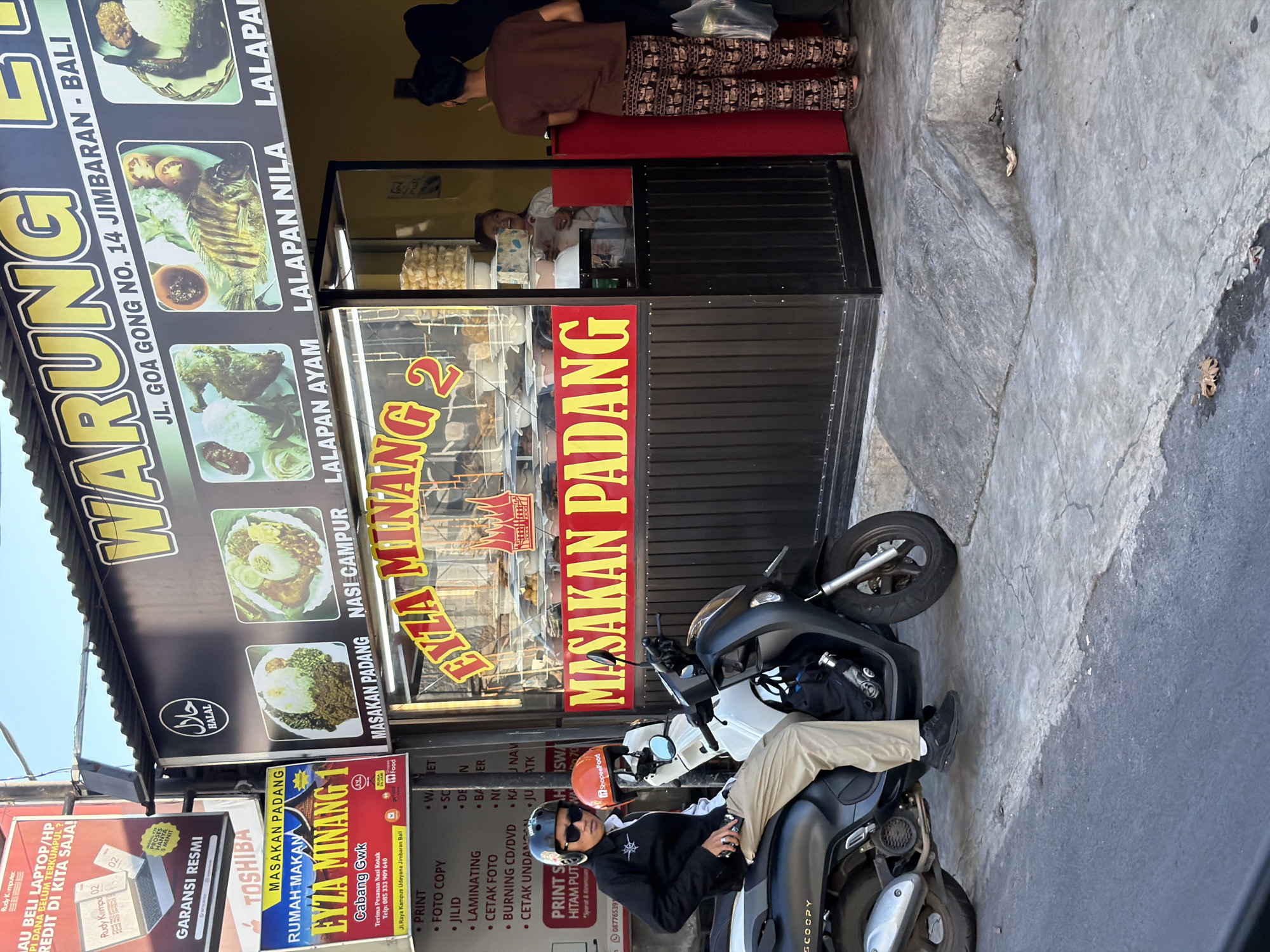 A wide shot captures the exterior of a food stall in Kuta Selatan, Indonesia, with a focus on a motorbike and its rider. The stall, identified by a sign reading "WARUNG EYZA MINANG 1", is a food establishment specializing in "Masakan Padang". The stall features a glass display showcasing various food items. Above the stall, a banner advertises different dishes, including "MASAKAN PADANG", "NASI CAMPUR", and "LALAPAN NILA" with accompanying images.
In the foreground, a person wearing a black jacket, beige pants, and a black helmet sits on a black and white motorbike. The motorbike is parked on a concrete surface. To the right of the stall, two individuals are visible. One, facing away from the camera and wearing a brown t-shirt and patterned pants, appears to be interacting with someone inside the stall. The second individual, partially obscured by the first, is wearing dark clothing. The overall environment is an outdoor setting with a clear sky, suggesting it is daytime. The ground surface is paved with concrete, and there are various advertisements and signs in the background.