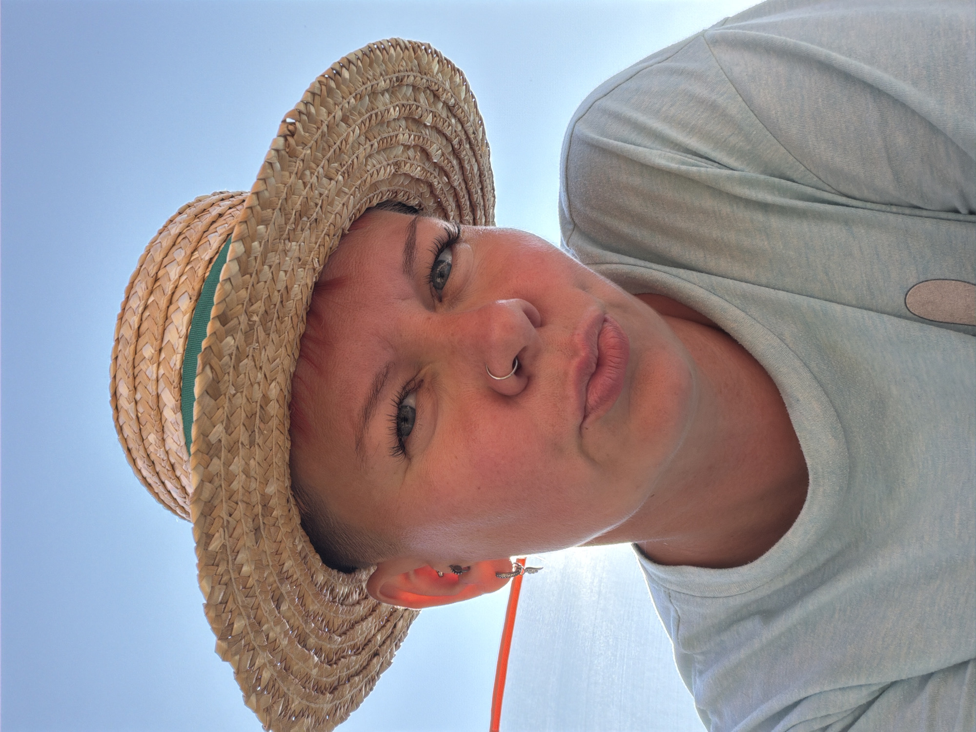 A close-up, low-angle selfie shows a person wearing a straw hat with a green band, a light blue t-shirt, and earrings. The person has short, pink-tinted hair, a nose ring, and is looking at the camera with pursed lips. The background is a clear blue sky, suggesting an outdoor setting. There are no identifiable landmarks or features that would indicate the specific location of Chaikovsky, Russia. The image appears to be taken on a bright, sunny day.