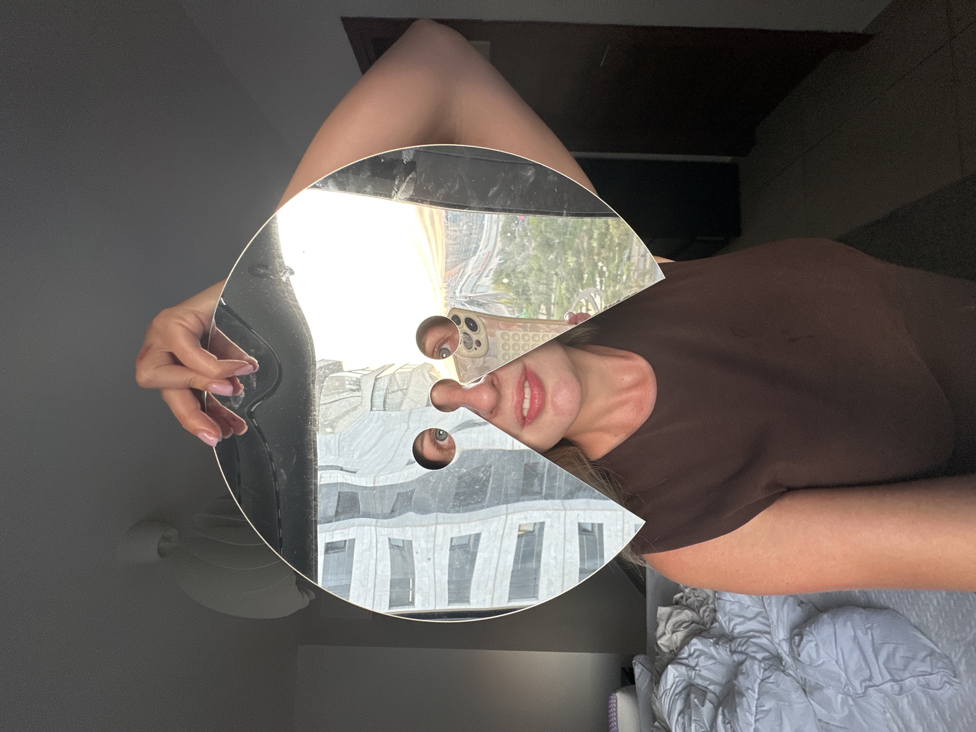 The image features a person, a woman, interacting with a large, circular mirror that has a segment removed, creating a pac-man-like shape. The woman is wearing a brown top. Her face is partially visible in the mirror, with her mouth open as if speaking or smiling. Her eyes are also reflected in the mirror, one clearly visible through a circular cutout in the mirror's surface, and another peeking through another smaller cutout. A smartphone, with a patterned case, is visible in the reflection, held in a way that suggests the woman is taking the photo or video.
The background reflected in the mirror shows a building with multiple windows, suggesting an urban or semi-urban setting. The lighting indicates it is daytime, likely with bright sunlight. The woman's posture and the way she is holding the mirror suggest a playful or curious activity, perhaps exploring her reflection or experimenting with the mirror's unique shape.
The scene is set indoors, with a bed covered in gray bedding visible in the lower right portion of the frame. The overall impression is a personal and informal moment captured through a creative use of a mirror. The location context provided is Petah Tikva, Israel. There is no visible text in the image.