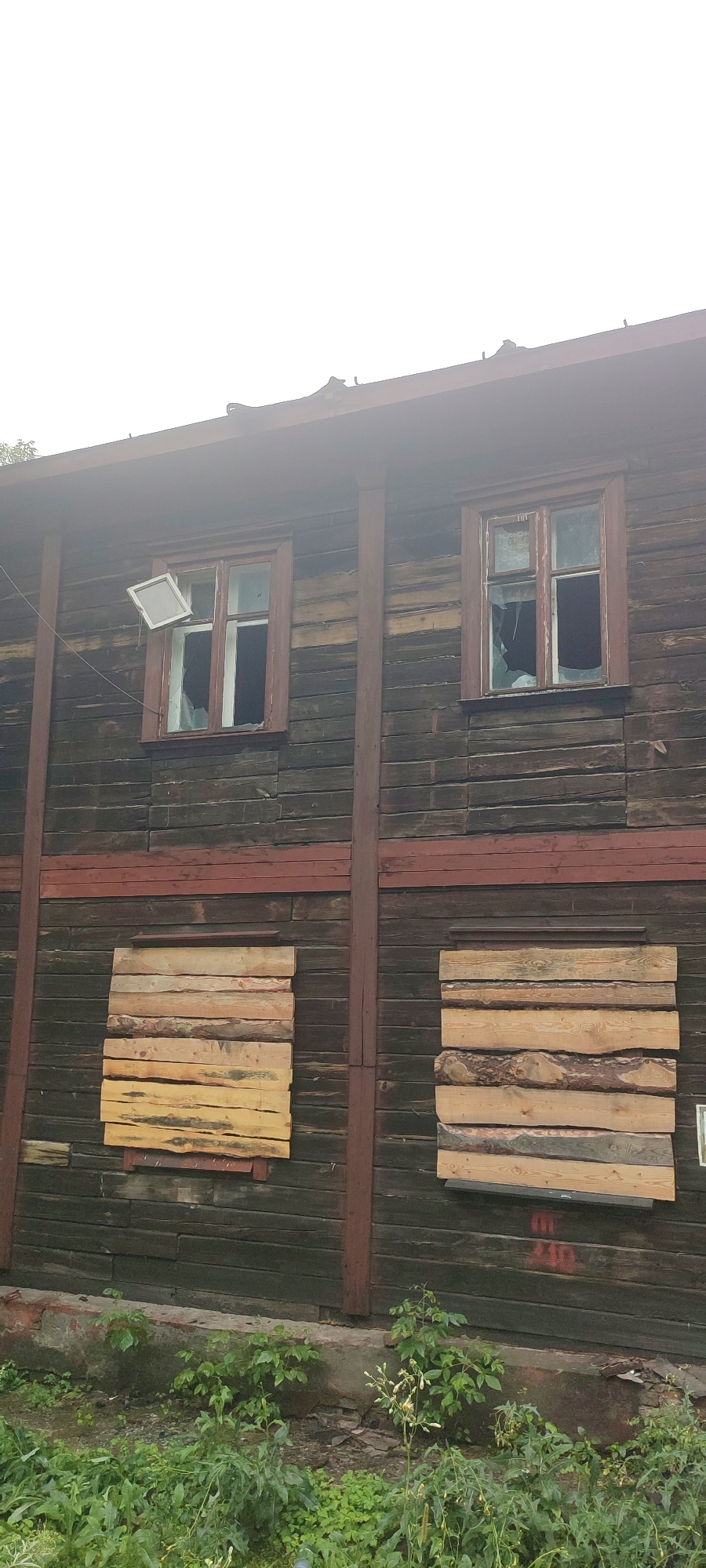 This is a vertical, full-shot photograph of the exterior of a dilapidated, two-story wooden building in Novoaltaysk, Russia. The building is made of dark, horizontal wooden planks, with vertical dark red wooden support beams. The upper story has two windows; the left window has a broken pane and a square, white panel leaning at an angle inside. The right window also has a broken pane, with dark fabric visible behind it. The lower story has two boarded-up windows. The left boarded-up window is covered with rough-hewn, pale yellow wooden planks arranged horizontally. The right boarded-up window is covered with similar, but rougher, pale yellow planks. Red graffiti, partially visible, is present on the lower section of the wooden wall between the two boarded-up windows. The ground in front of the building is covered with green vegetation, including grass, weeds, and some flowering plants. The foundation of the building is made of concrete and brick, showing signs of disrepair. The sky is overcast and white.