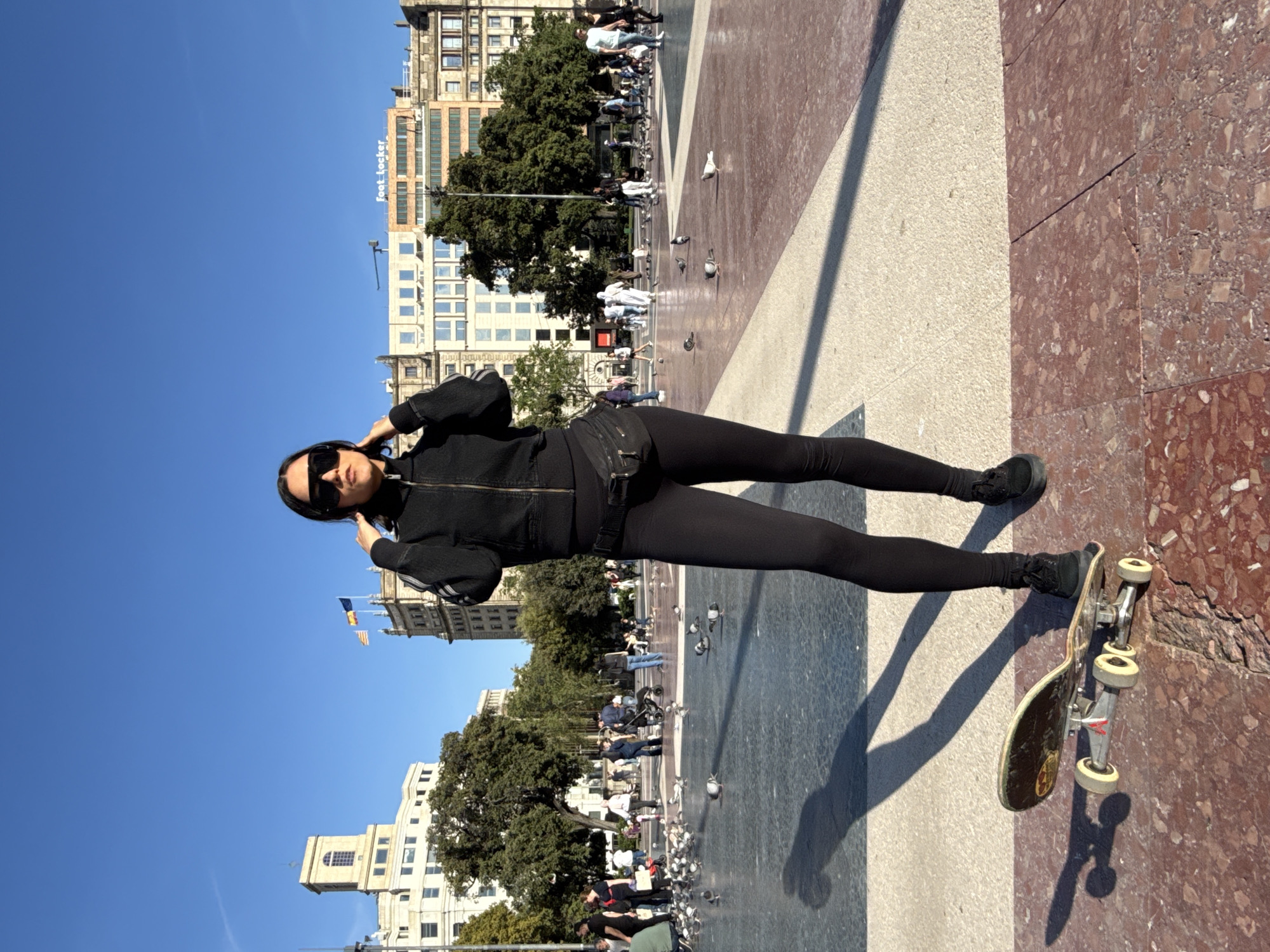 A young woman in a black jacket, leggings, and sunglasses stands with a skateboard in Plaça de Catalunya, Barcelona, Spain. The square is paved in a geometric pattern and populated by pedestrians and pigeons. Buildings are visible in the background under a clear blue sky.