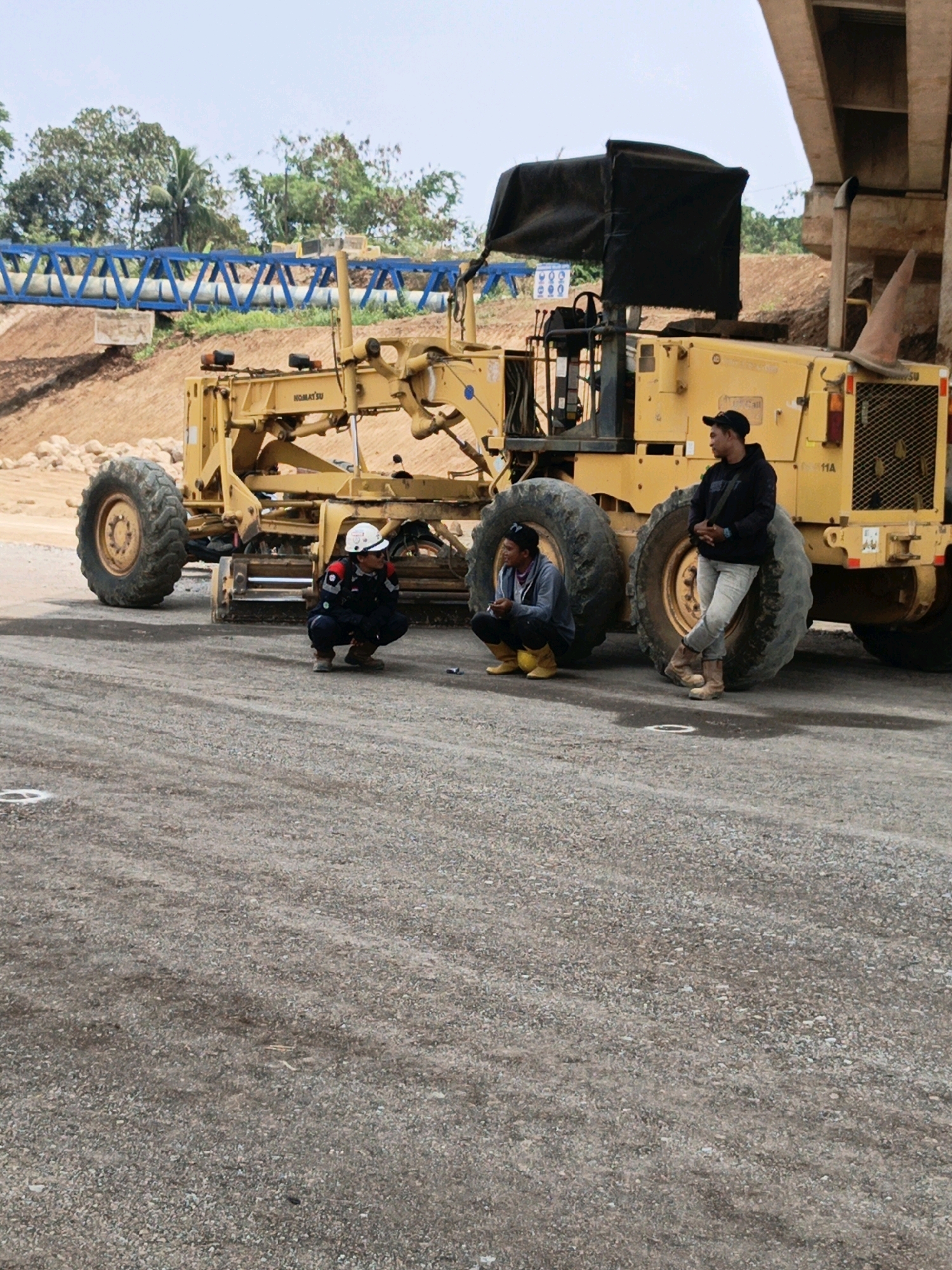 In Cibadak, Indonesia, three men are gathered near a large yellow construction grader. The man on the far left, wearing a white hard hat, dark work jacket, and blue pants, is squatting. To his right, another man in a grey hooded sweatshirt and yellow boots is also squatting. Leaning against one of the grader's massive tires on the right is a third man wearing a black hoodie, dark baseball cap, and light-colored pants. The grader, a Komatsu, is parked on a gravel surface. In the background, a blue bridge structure is visible, along with trees and a dirt embankment. The sky is overcast.