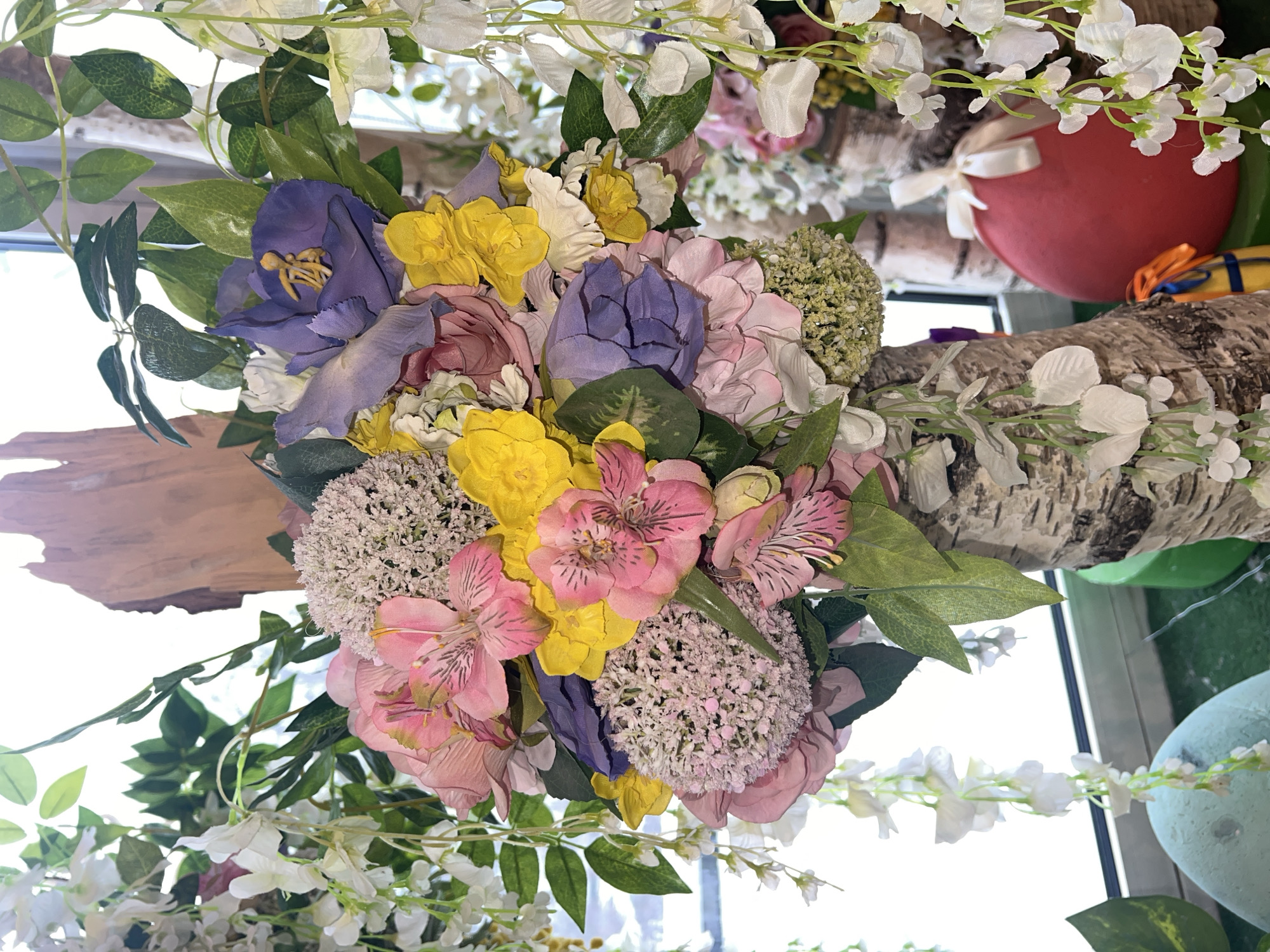 This image captures a vibrant, overhead decorative display, primarily featuring an artificial floral arrangement. The central focus is a large, colorful bouquet composed of various types of fabric flowers. Prominent blooms include bright yellow ruffled flowers, distinct purple-blue irises with yellow stamens, and pink alstroemeria (Peruvian lilies) with characteristic striped petals. These are complemented by clusters of light pink or mauve hydrangea-like blossoms and numerous small white flowers, some forming cascading garlands. Green foliage and leaves are interwoven throughout, providing contrast and volume.
The floral arrangement is integrated with other decorative elements, notably sections of birch logs or branches, which appear to be part of the structure or simply adding a rustic touch to the decor. In the upper right background, a bright red heart-shaped object, tied with a white ribbon, is visible, suggesting a theme of love or celebration. Further into the background, indistinct colorful shapes are present, including a light blue-green spherical item in the bottom right.
The setting appears to be indoors, likely a commercial or event space, given the elaborate and permanent-looking nature of the decor. Bright, natural light from what seems to be a window or skylight illuminates the scene, indicating daytime. There are no people visible, nor any specific activity taking place; the image is a static portrayal of the decorative setup. No visible text is present. The overall impression conveyed is one of joy, beauty, and a festive atmosphere.