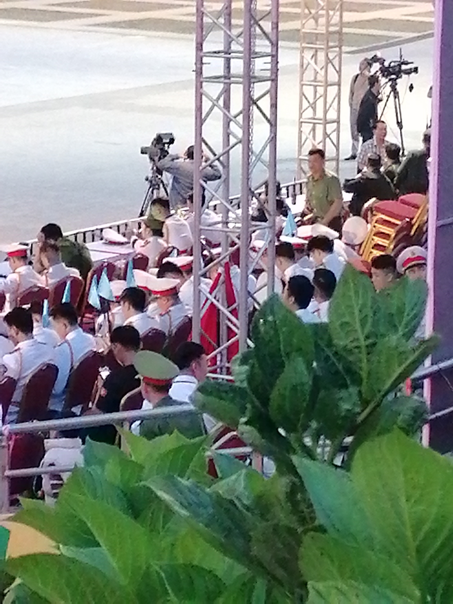 The image depicts a gathering of people, possibly at an outdoor event or ceremony, in Da Lat, Vietnam. In the foreground, large green leaves obscure the lower portion of the scene. Behind the foliage, a crowd of individuals, many wearing white shirts and hats, are seated in rows of chairs. Several people in the crowd are wearing red and white striped hats.
Further into the background, a man in a green uniform and a hat is visible. To the left, a video camera on a tripod is being operated by a person. Behind the camera operator, a group of people, some appearing to be in uniform, are gathered. Construction scaffolding structures are present, suggesting the event may be taking place in a temporary setup or an area under construction. Red chairs are stacked in the background. The overall setting appears to be an open area, likely a public space in Da Lat, given the context provided. The lighting suggests it is daytime.