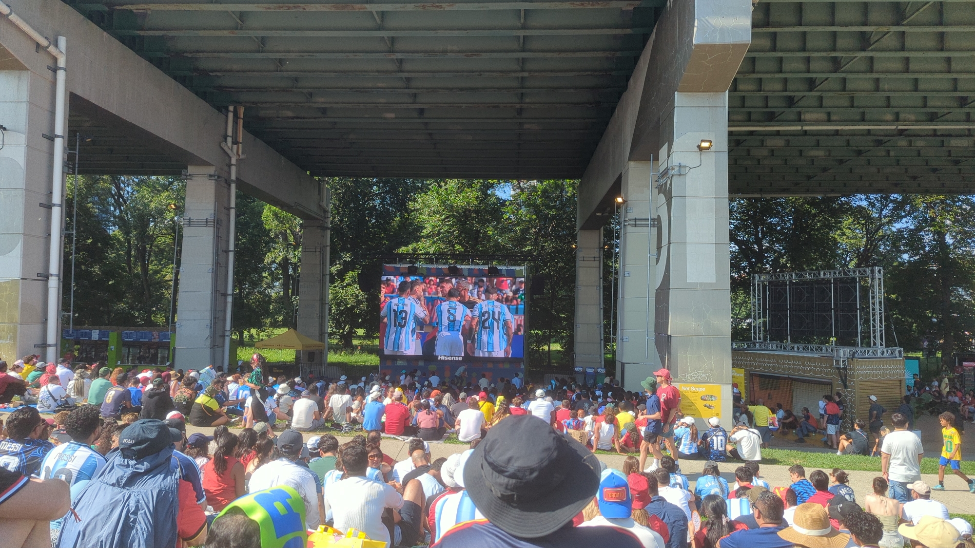 A large crowd of people are gathered outdoors, seated on the ground and facing a large screen. The screen displays a video of soccer players in light blue and white striped jerseys. The gathering appears to be taking place under a large overpass or bridge structure with concrete pillars. Behind the screen, trees and a bright blue sky are visible. To the right of the screen, there is a smaller setup that looks like a stage or display area. The crowd is diverse in age and is dressed casually, with many wearing soccer-themed apparel. The overall atmosphere suggests a public viewing event for a soccer match, possibly in a park or open public space within the city of Toronto, Canada, as suggested by the presence of a yellow tent with text that appears to be in Spanish, common in Toronto's diverse communities. The lighting indicates it is daytime.