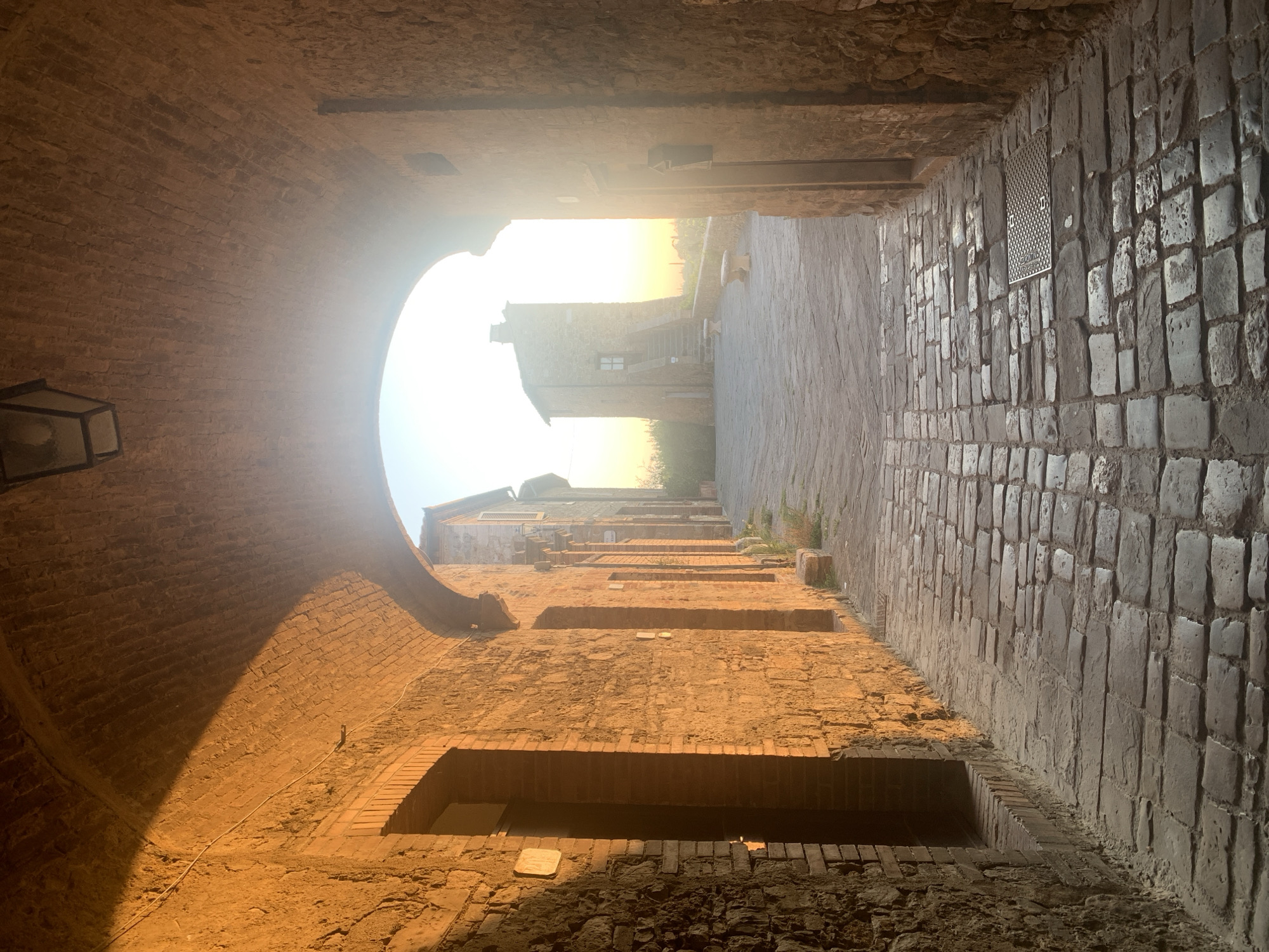 The image depicts a stone-paved alleyway in San Quirico d'Orcia, Italy, likely in the historic center. The scene is illuminated by the warm glow of the setting sun, which casts long shadows and highlights the textured brick and stone architecture. An arched passageway frames the view, leading to a small piazza. In the distance, a stone tower with an external staircase stands prominently against a bright sky. Stone benches are visible in the piazza. The alley floor is made of irregular cobblestones, and a metal grate is embedded in the pavement. The walls of the buildings are constructed from rough stone and brick, showing signs of age. The overall atmosphere is tranquil and historic.