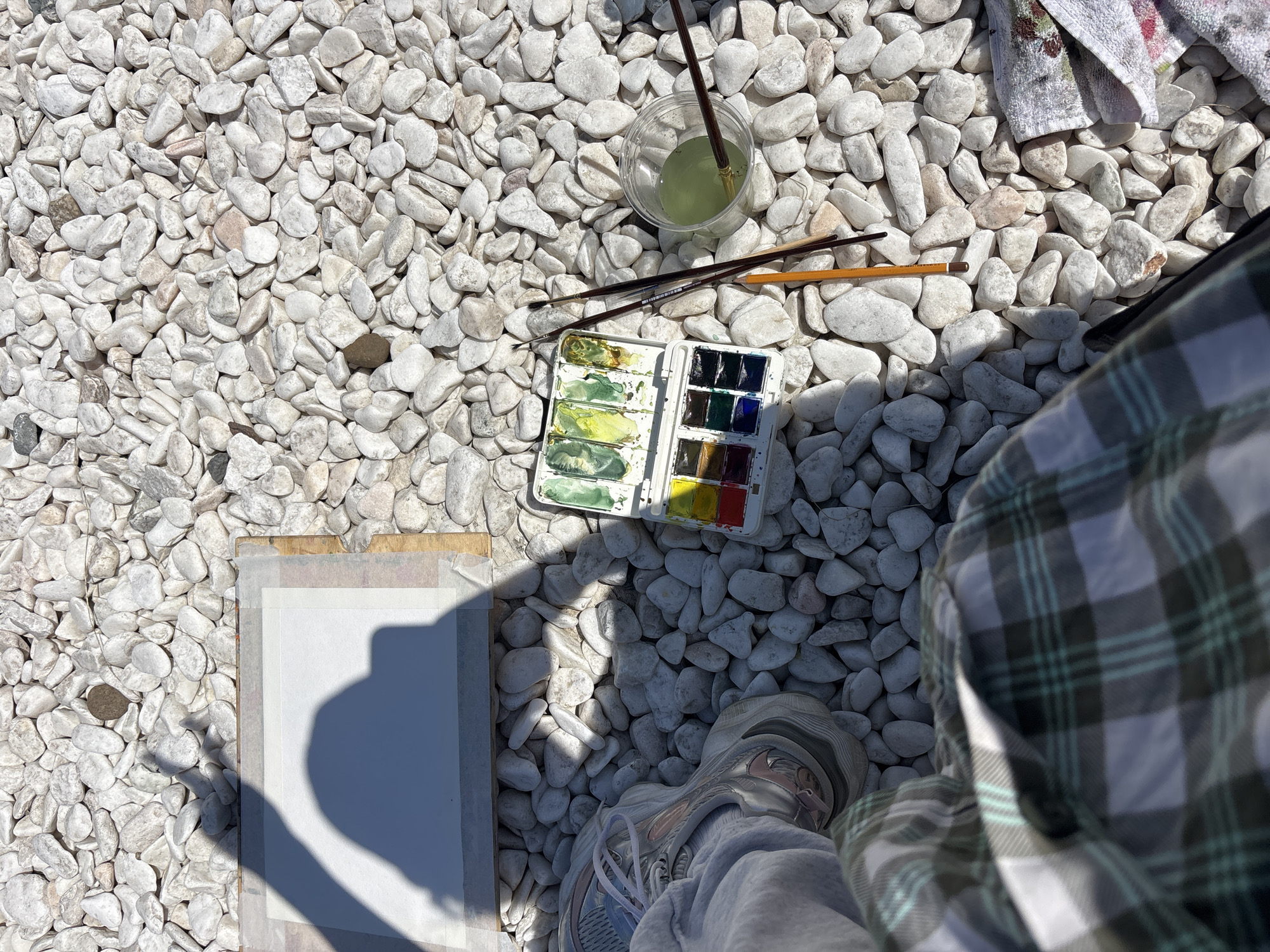 An overhead, high-angle shot captures an outdoor scene featuring art supplies laid out on a bed of bright white, smooth pebbles. Strong, distinct shadows are cast by the high sun, suggesting clear weather during midday or early afternoon in Дрокино, Russia.
In the center of the frame, an open white watercolor palette displays a range of colors, including vibrant greens, yellows, blues, and reds. Several paintbrushes are scattered nearby, one clearly showing the brand "Koh-i-noor." Above the palette, a clear plastic cup holds murky, greenish water, with a paintbrush soaking inside. A folded, stained white towel is visible in the upper right corner.
In the bottom left, a sheet of white paper, likely watercolor paper, is taped onto a wooden board. A prominent, elongated shadow of a person, presumably the artist, falls across this paper. The artist's presence is further indicated by a foot clad in a light-colored athletic shoe and grey sweatpants, visible in the bottom left, and a portion of a plaid shirt (green, black, white pattern) and grey sweatpants on the right side of the frame.
The overall scene depicts a person engaged in or preparing for an outdoor watercolor painting session, using the pebble-covered ground as a creative workspace.