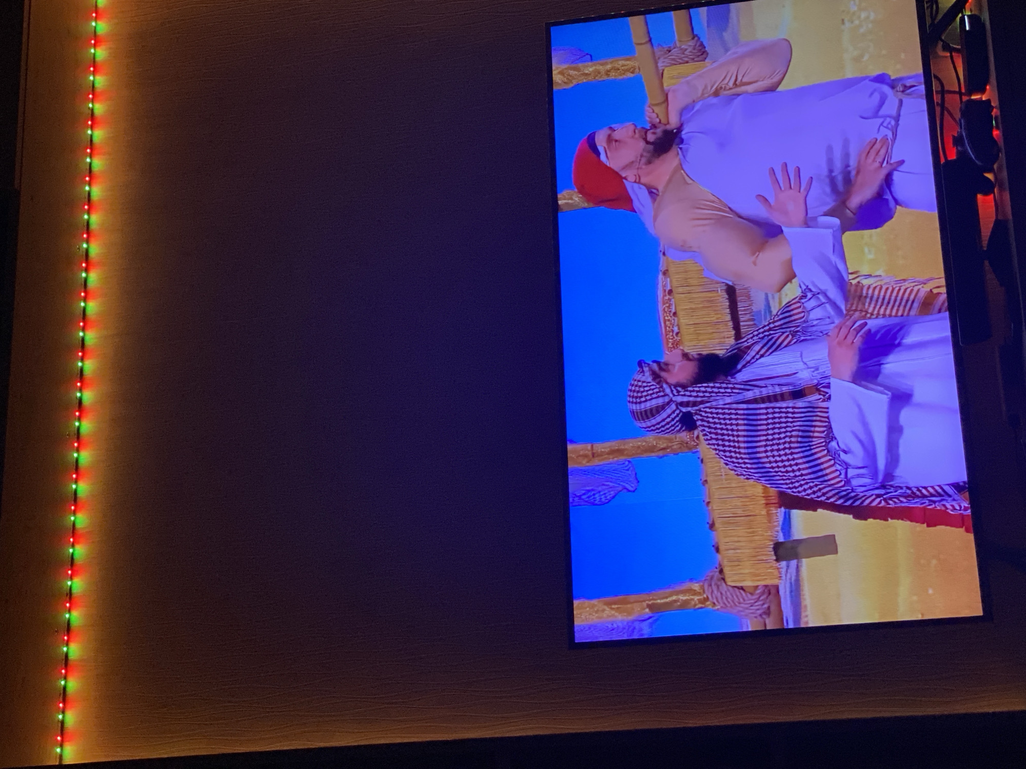 The image displays a television screen showing a scene with two individuals dressed in traditional Middle Eastern attire. The person on the left wears a keffiyeh and a white robe, while the person on the right has a red head covering and a light-colored tunic. They appear to be interacting in a desert-like setting depicted on the screen. In the foreground, below the television, is a dark surface with some electronic devices, including what looks like a video game controller. Above the television, a string of red and green LED lights is attached to a wall, illuminating the upper portion of the image. The overall environment suggests an indoor setting where a television is being watched. The provided information does not specify a location within Chișinău, Moldova.