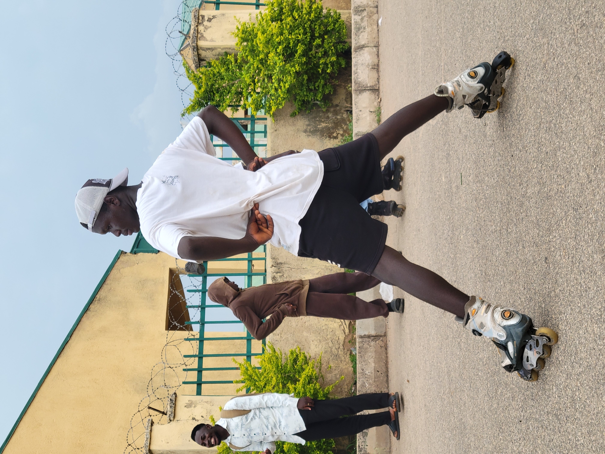 A group of three men is standing on a paved surface, likely a road or pathway. The man closest to the camera is wearing a white t-shirt, black shorts, a cap, and inline skates. His legs are spread wide, and he is holding his hands on his lower back. Behind him, a man wearing a brown hoodie and pants is standing with his hands on his lower back. Further behind and slightly to the side, another man in a patterned white shirt and dark pants is standing and smiling. The background includes a light yellow building with green trim and barbed wire, a green fence, and some greenery. Based on the building style and landscape, the location may be in Jos, Nigeria.