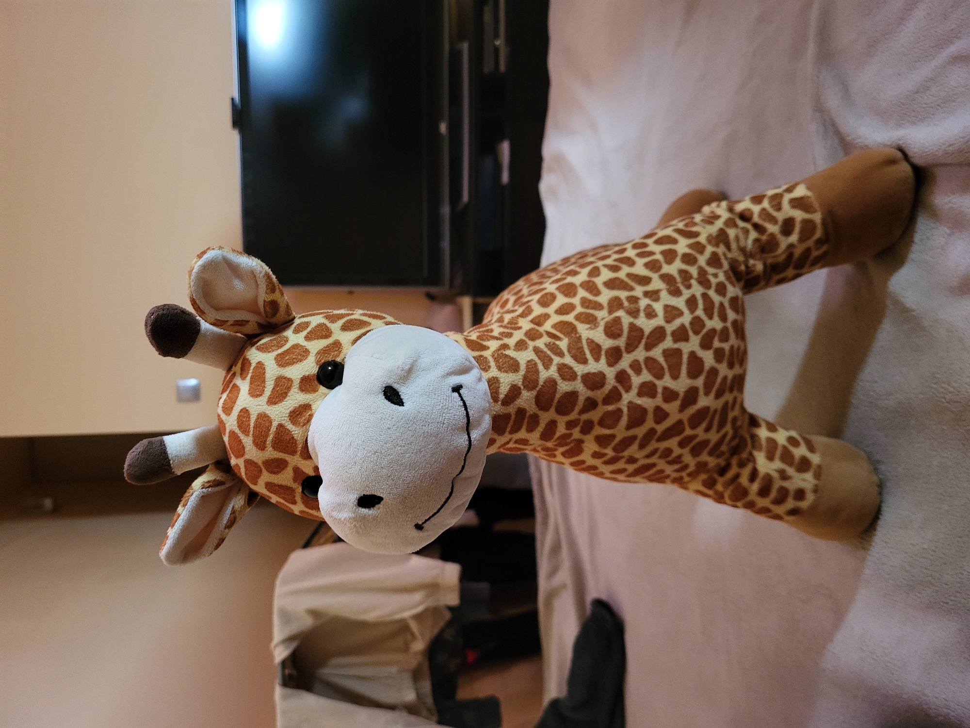 A stuffed giraffe toy sits on a bed. The giraffe is primarily light brown with darker brown spots. It has a white face with black button eyes and a stitched smile. Two brown horns with white tips protrude from its head, and its ears are lined with white. The giraffe is positioned facing forward, with its front legs slightly apart. The bed is covered with a light-colored, textured fabric. In the background, a dark television screen is visible, mounted on a dark stand. To the left of the television, there is a rack with clothing draped over it. The lighting in the scene appears to be artificial, casting soft shadows. The location is unknown.