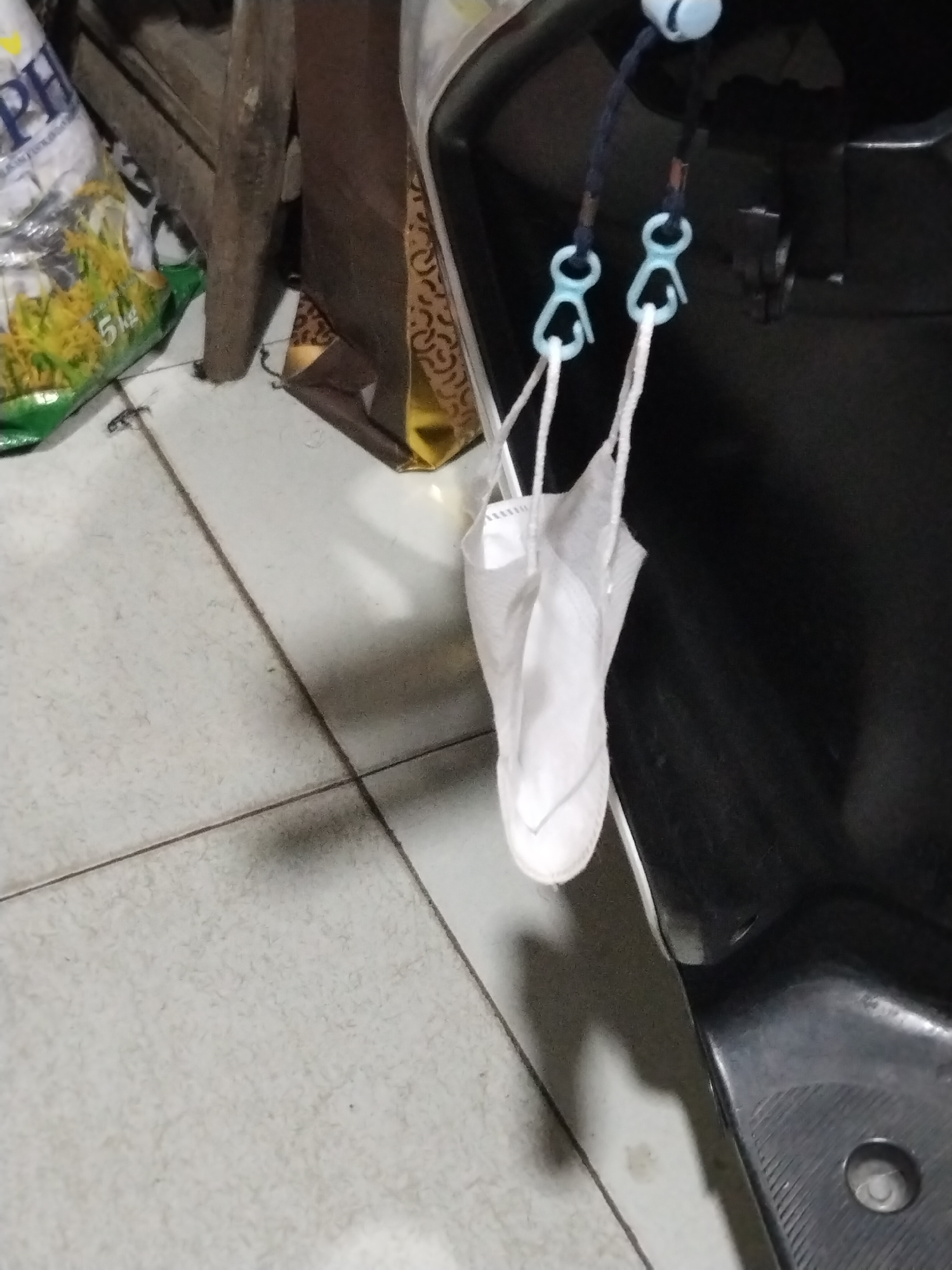 An indoor scene shows a white disposable mask hanging by its ear loops from a dark blue cord with two light blue plastic clasps. The cord is attached to a light blue cylindrical object, likely a bead or stopper, which is affixed to a dark surface. To the left of the hanging mask, partially visible, is a green and white bag with the letters "PH" and the number "5kg" printed on it. Behind the mask and cord, the dark surface appears to be part of a motorcycle or scooter. The floor is tiled with light-colored square tiles and has dark grout lines. The lighting suggests an indoor environment, possibly a garage or storage area. The overall impression is one of everyday objects stored in a functional space.