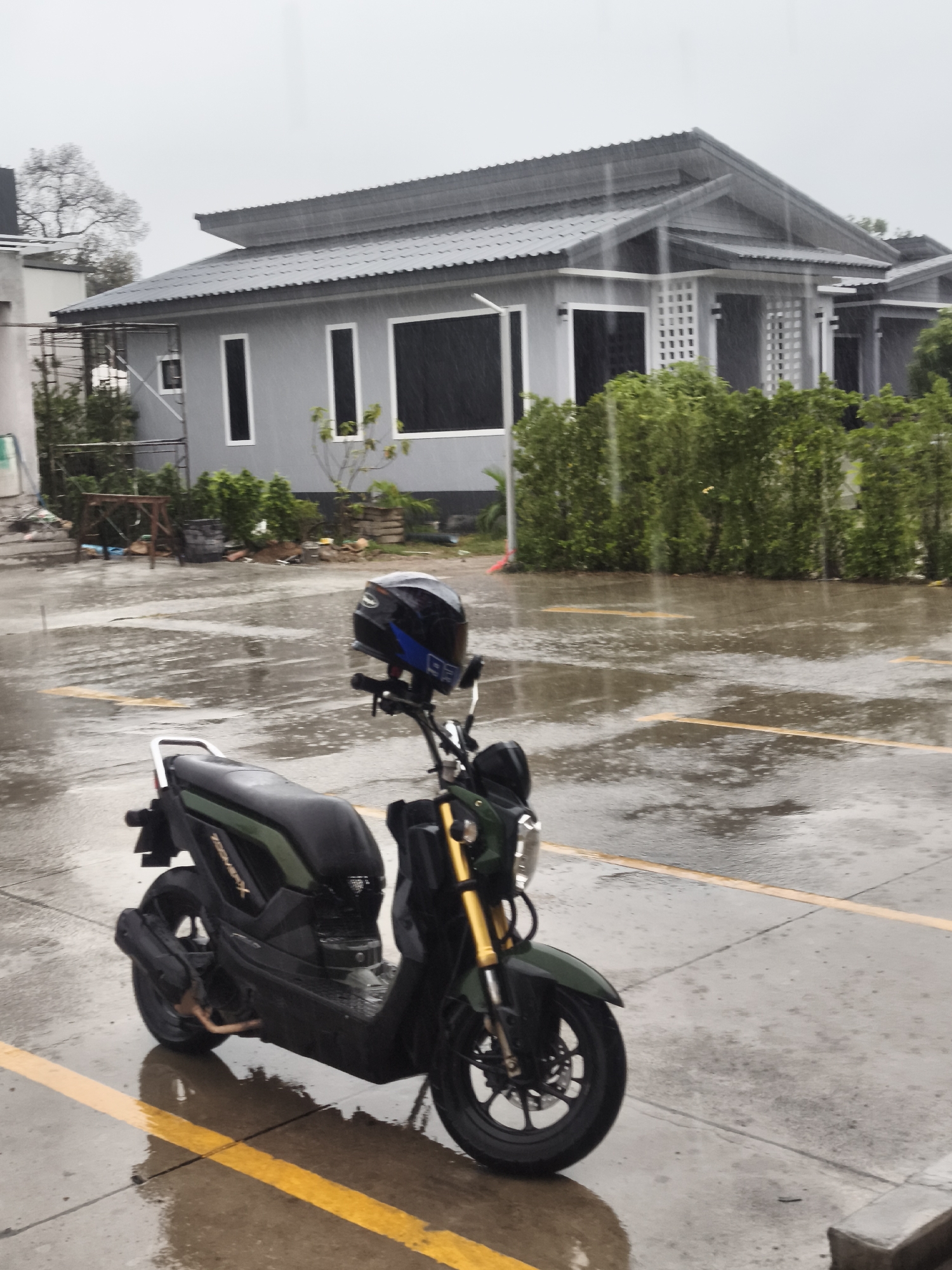 In the city of Ko Samui District, Thailand, during a rain shower, a dark green scooter with a black seat is parked on a wet paved surface marked with yellow lines. A black motorcycle helmet with blue accents sits on the handlebars of the scooter. In the background, several modern-style gray houses with dark roofs are visible, surrounded by lush green bushes. The sky is overcast and a steady rain is falling, evident by the streaks in the air and the reflections on the wet ground. Construction scaffolding is visible near one of the houses.