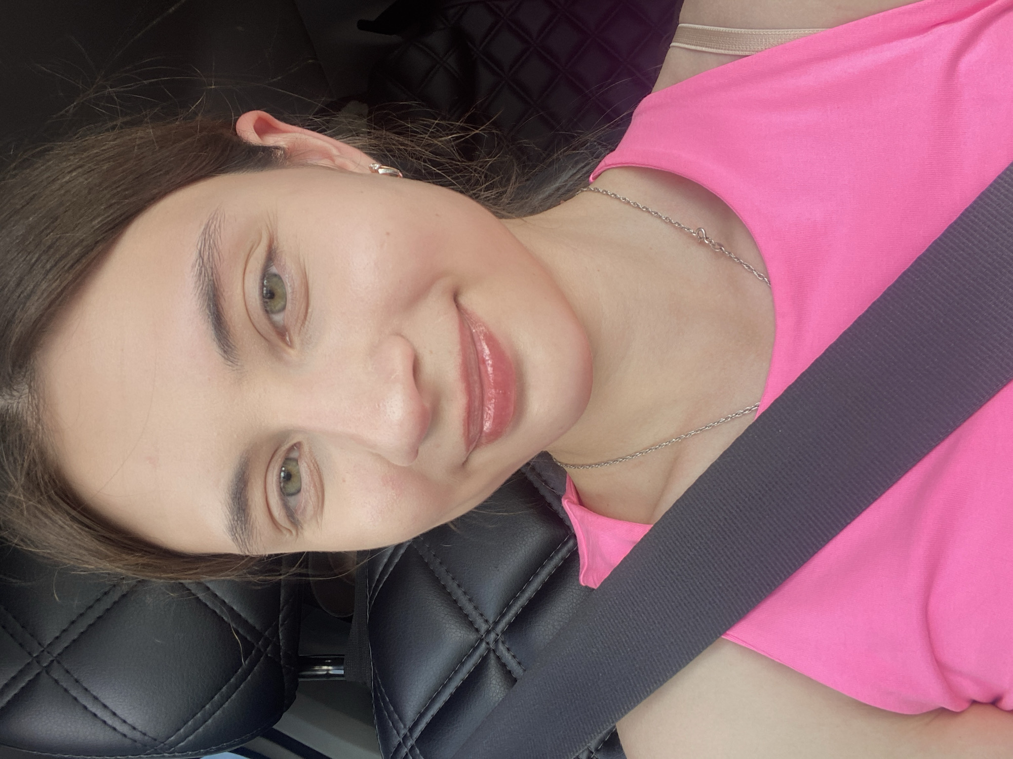 The image shows a close-up, front-facing selfie of a young woman inside a vehicle. She has fair skin, green eyes, and brown hair pulled back. She is wearing a bright pink tank top and a silver necklace. A grey seatbelt is diagonally positioned across her chest. The background includes a black, quilted car seat and her hair. The setting is consistent with being inside a car, likely in the city of Kamkoye, Russia, given the implied context. There are no other people or significant environmental details visible.