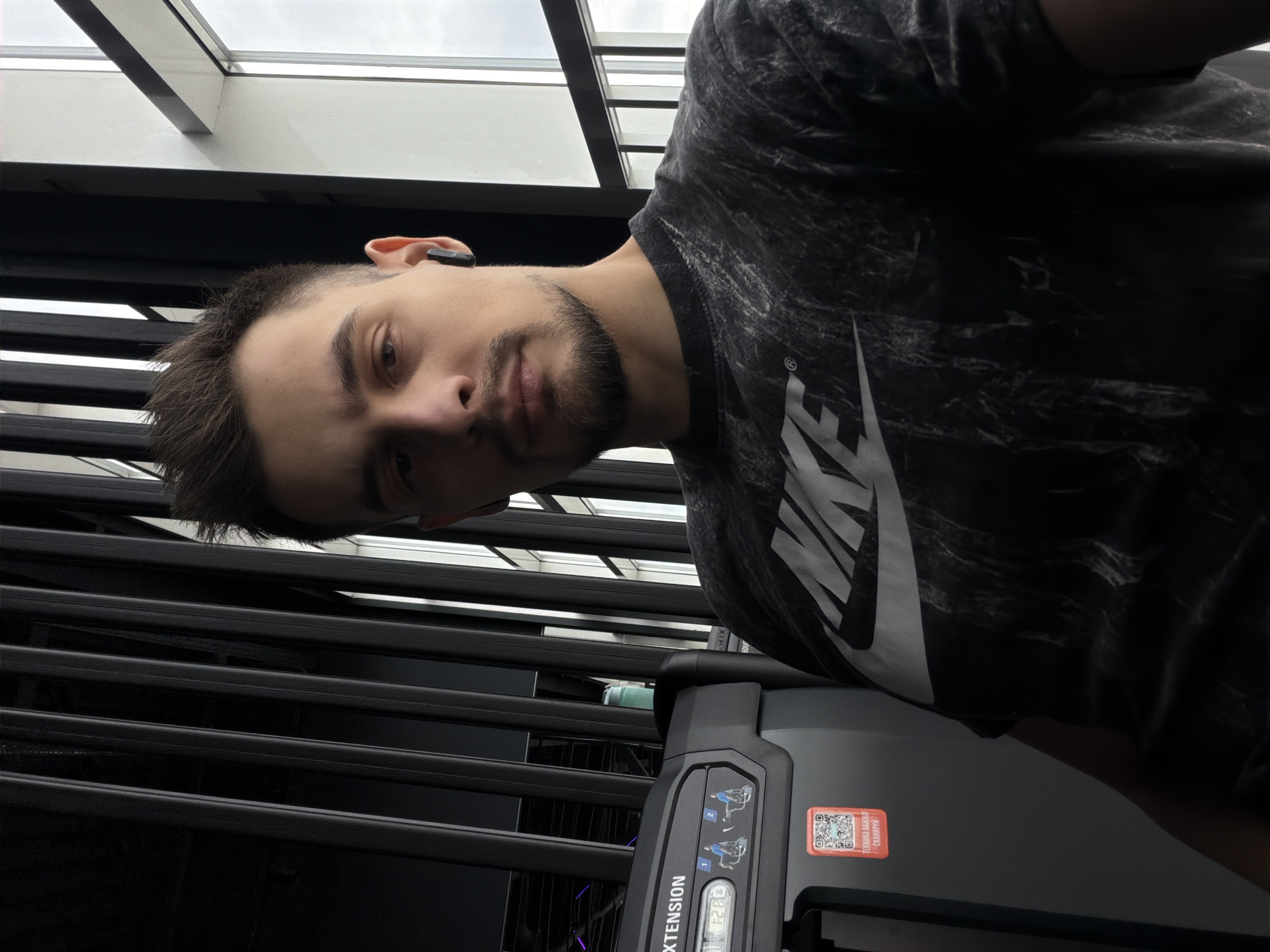 A close-up, tilted indoor shot depicts a man with short brown hair and a beard, wearing a dark t-shirt with a white Nike logo. He is looking directly at the camera with a neutral expression. The man is wearing a small, dark-colored device in his left ear. He appears to be in a gym, given the exercise equipment visible in the lower portion of the image. The equipment is gray and black with control panels and diagrams. Black horizontal bars are visible in the background. Above the bars, there's a section of a window with a cloudy sky visible through it. The image contains a QR code on the equipment. The location is the city of Krasnoyarsk, Russia.