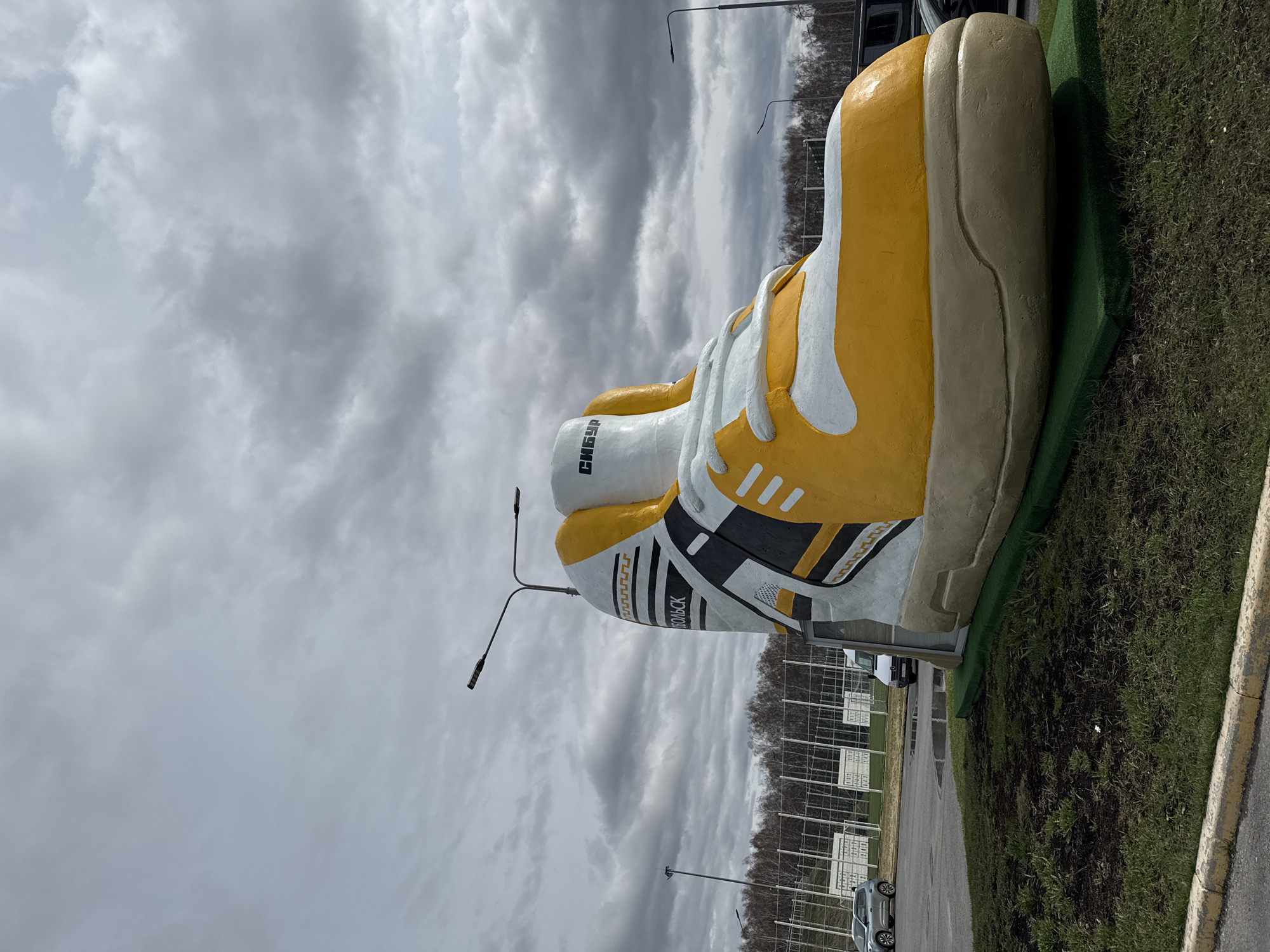 A large, painted concrete shoe sculpture is located on a patch of grass near a road in Tobolsk, Russia. The shoe is yellow and white with black detailing and has the words "СИБУР ТОБОЛЬСК" painted on its side. In the background are parked cars and a grey cloudy sky.