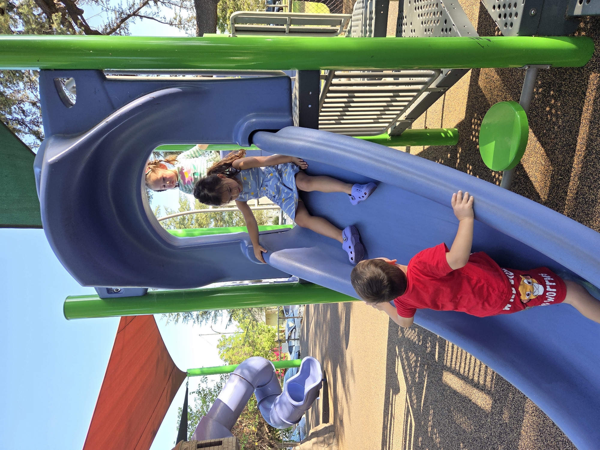 In a sunny outdoor playground in Glendale, California, three young children are interacting with a blue and green plastic slide. The slide is part of a larger playground structure with purple tube slides and a red sun shade. A young boy in a red t-shirt and red shorts with a Lion King character is at the bottom of the slide, looking up. A young girl in a blue and white patterned dress is sliding down. Another child, a girl with blonde pigtails, is at the top of the slide, looking down. The ground is covered with a brown rubberized surface with dark speckles. Sunlight casts shadows on the ground.