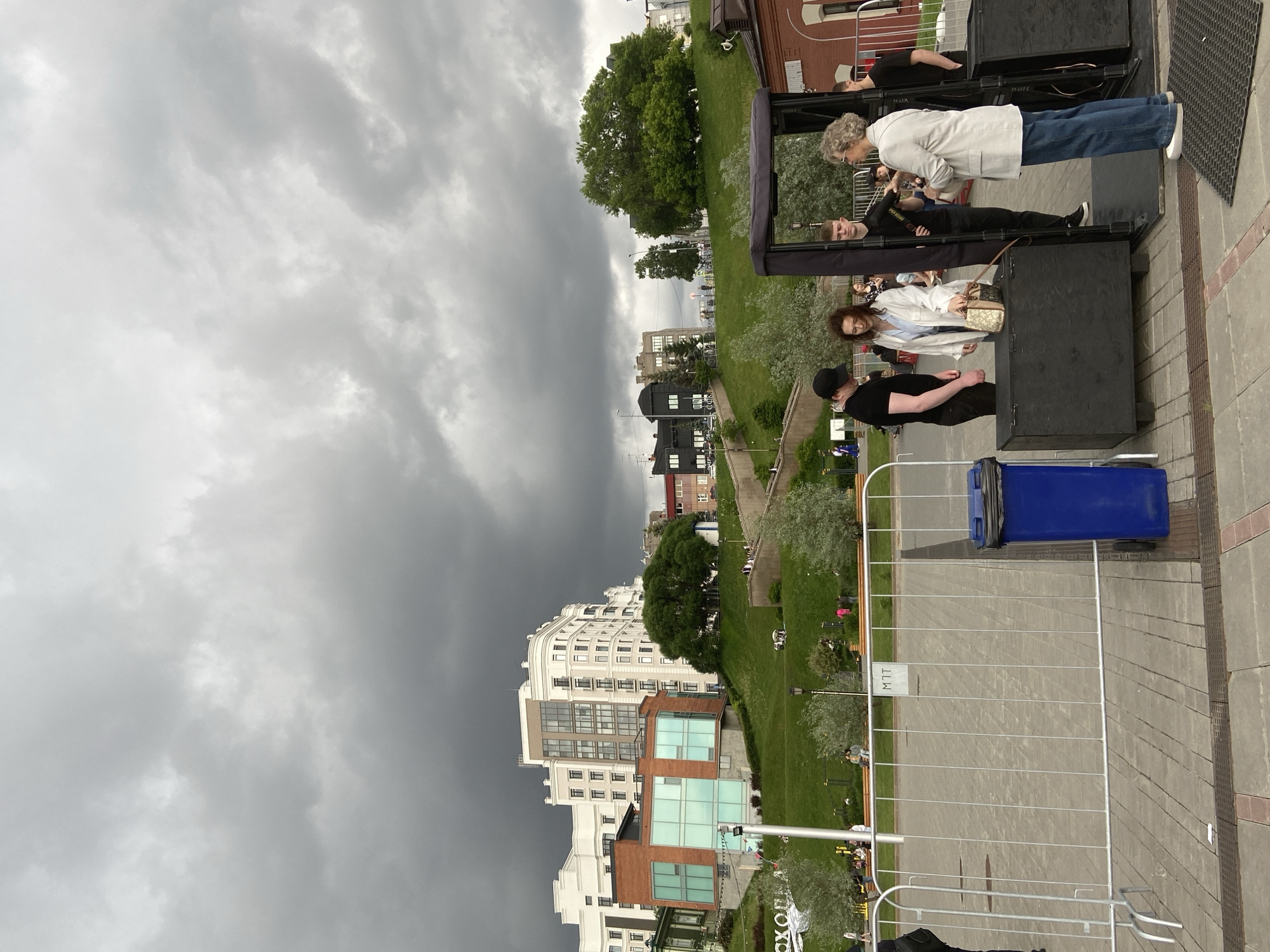 The image captures an outdoor urban scene under a heavily overcast sky, hinting at potentially stormy weather. The setting appears to be a public square or park, with paved areas in the foreground leading to grassy slopes dotted with trees. Modern buildings, some multi-story and white, others featuring brick and glass facades, line the horizon in the background. The provided context indicates the location is Tyumen, Russia.
In the foreground, a group of people is gathered around what appear to be black stage components or large speakers. Among them, a woman with short, curly grey hair wears a light-colored jacket and blue jeans, standing next to a man with a camera strap around his neck. Another woman with curly brown hair, dressed in a white shirt, holds a small patterned handbag. A man in a black t-shirt and black cap stands nearby. Further back, another person leans against a structure. The arrangement suggests an outdoor event, possibly a performance or public gathering, with crowd control barriers separating the foreground area from a wider paved space.
A large blue wheeled trash bin is visible to the right, next to the metal barriers. The barriers themselves extend across the foreground, defining a clear space. On one of the white signs attached to the barriers, the letters "MTT" are visible. In the mid-ground, more people are casually distributed across the grassy areas, some sitting on benches or walking. The overall impression is an active public space on a cool, cloudy day.