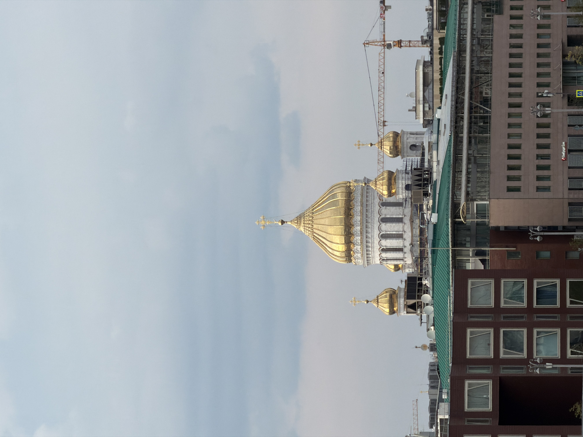 The image shows a view of the Cathedral of Christ the Saviour in Moscow, Russia, from a distance. The cathedral's iconic golden domes are visible, along with several smaller domes. The cathedral is surrounded by other buildings, including office buildings and residential complexes. A construction crane is also visible in the background, suggesting ongoing development or renovation in the area. The sky is overcast with clouds. The foreground features the upper portions of several modern buildings, characterized by their rectangular shapes and numerous windows. The building on the left is dark red, while the building in the center is topped with a green roof. The building in the foreground right is a light brown color with many rectangular windows.