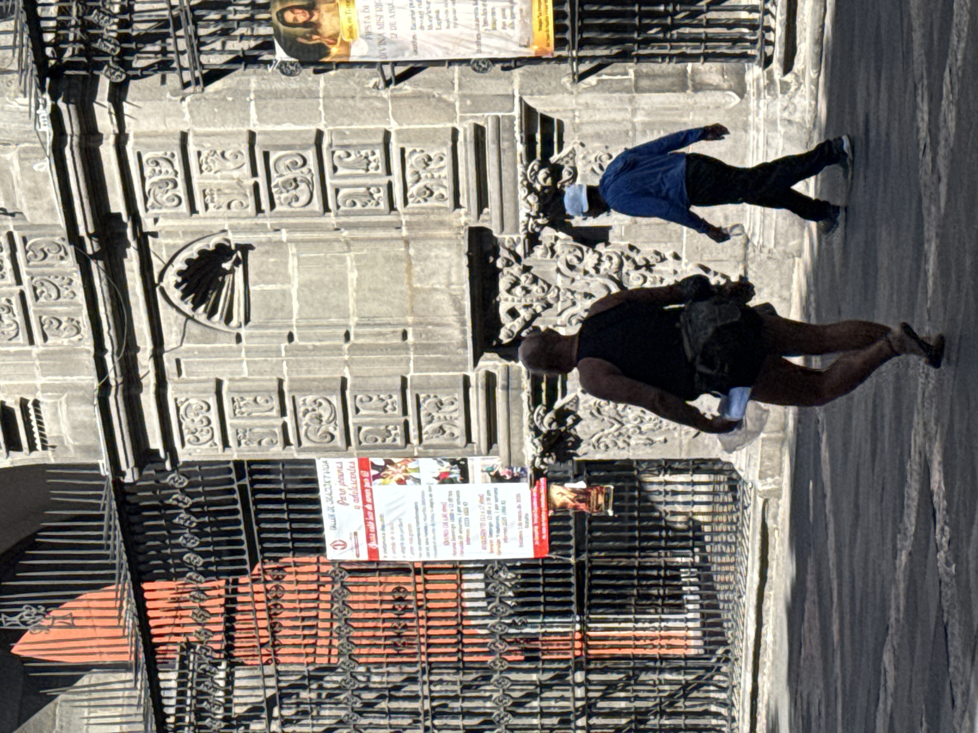 The image captures a street scene in Puebla, Mexico, adjacent to a historic building with detailed stonework. Two individuals are prominently featured: one wearing a blue shirt and a face mask, while the other is clad in a black tank top and carrying a bag. Both are in motion. A poster labeled "TALLER DE ORACION Y VIDA" is affixed to a barred window. A sign reading "FIESTA DE DIVINA MISERIC" is also visible. The environment consists of a stone building facade, possibly a church or governmental structure, with ornate details. The street surface is paved, reflecting sunlight.
