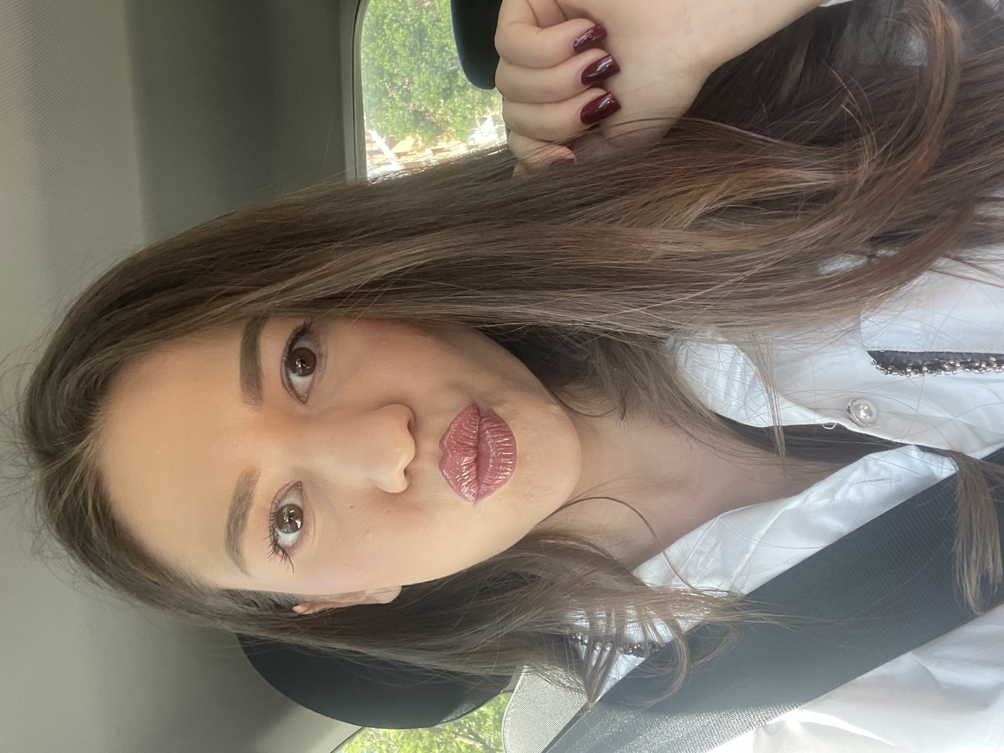 A woman with light brown hair and brown eyes is in a car. She has red nail polish and is wearing a white blouse and a black seatbelt. The location is unknown, and there is no indication of being in Sofia, Bulgaria.